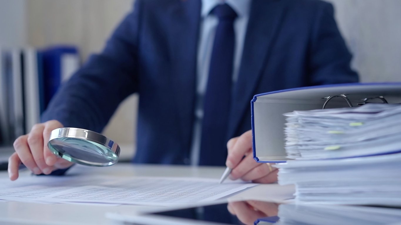 Person in a business suit examining documents with a magnifying glass at a desk.