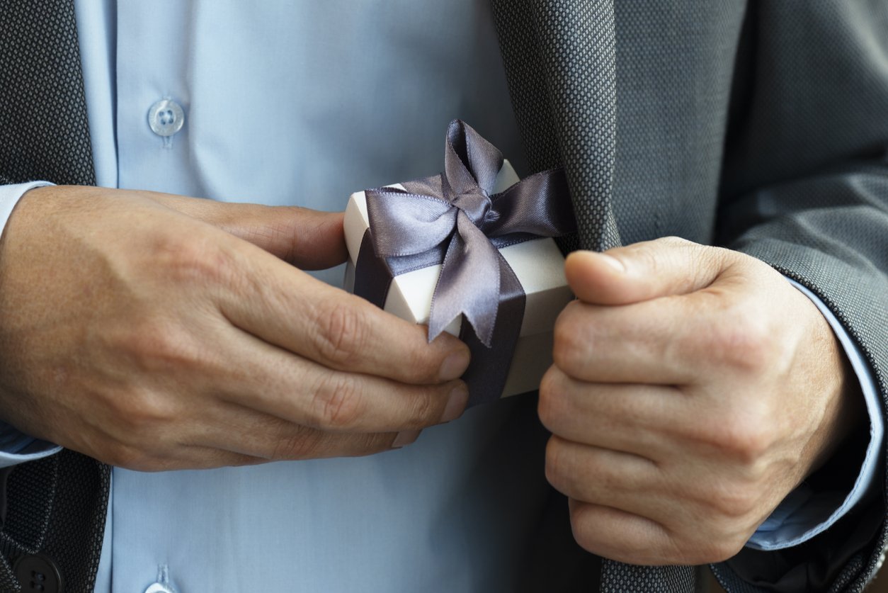 A person in a suit is holding a small gift box wrapped with a dark satin ribbon and bow.