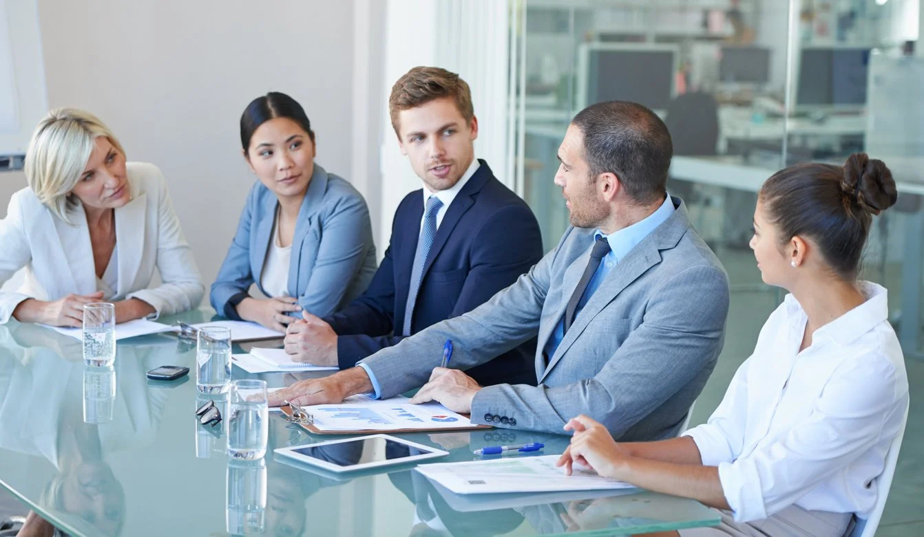 Five professionals engaged in a discussion at a business meeting in a conference room.