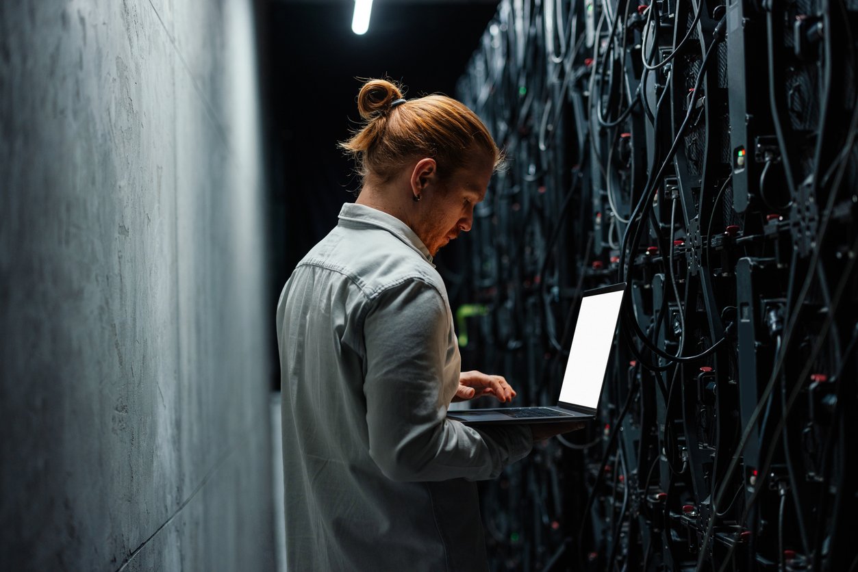 A man with red hair tied in a bun wearing a light-colored shirt using a laptop in a server room with racks of servers and cabling.