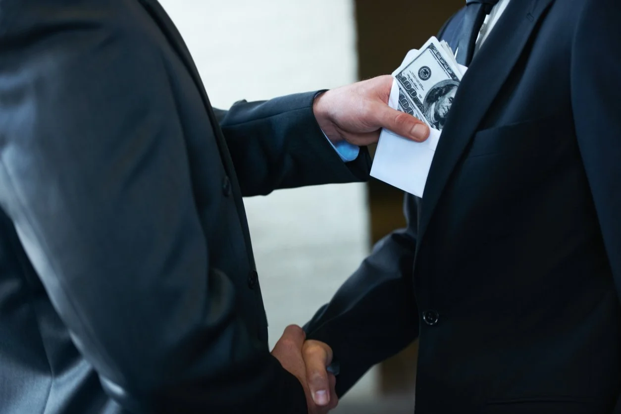 Two men in business suits shaking hands, one of them holding a stack of hundred-dollar bills in an envelope.