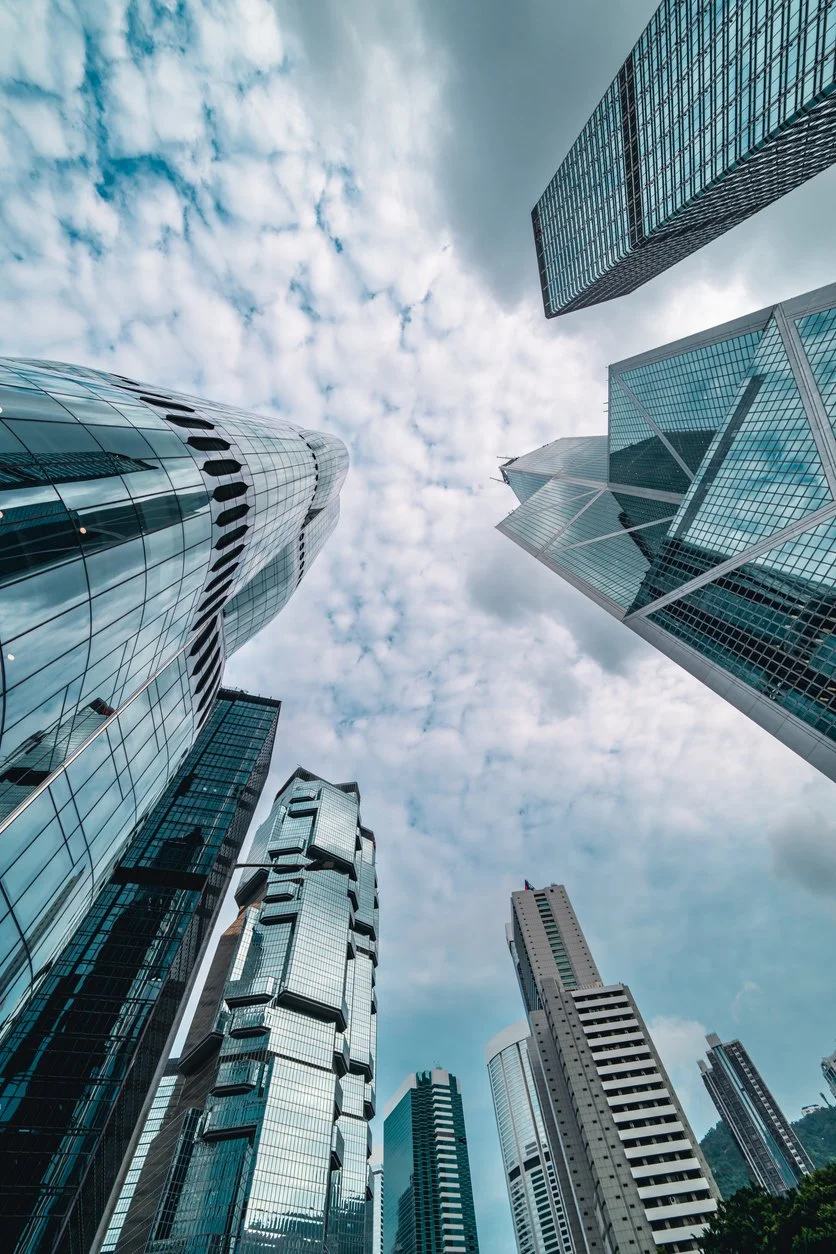 Looking up at tall modern skyscrapers with glass facades in a city skyline on a partly cloudy day.
