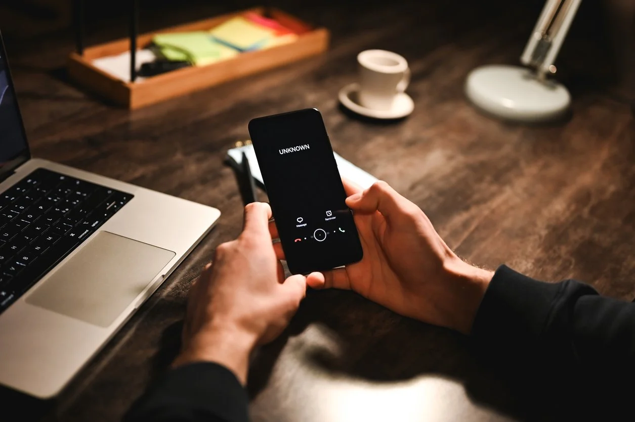 Person holding a phone during a call with 'UNKNOWN' on the screen, on a wooden desk with a laptop, coffee cup, desk lamp, and a wooden tray with colorful sticky notes in the background.