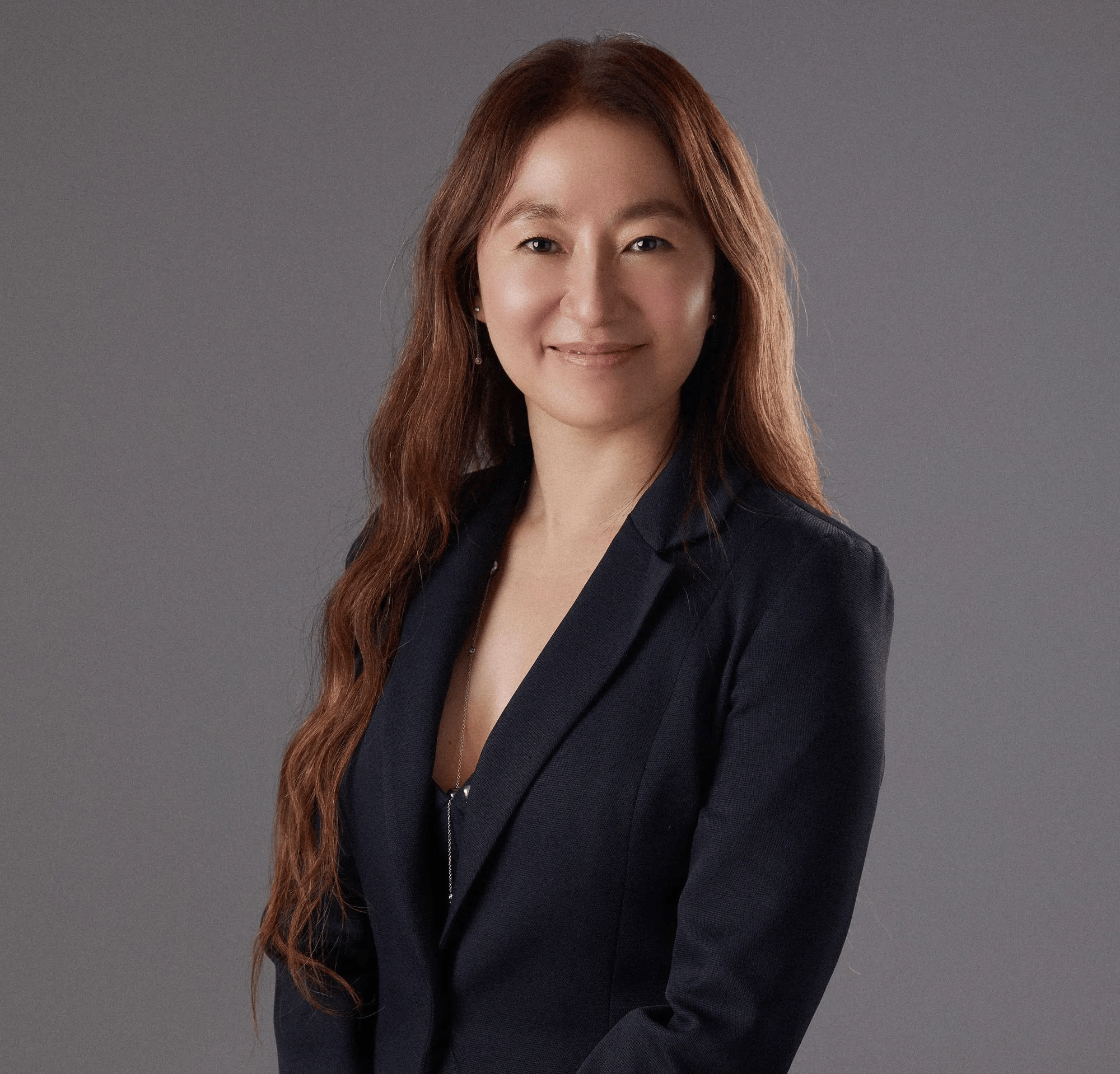 Portrait of a woman with long, wavy, auburn hair wearing a dark blazer against a gray background.