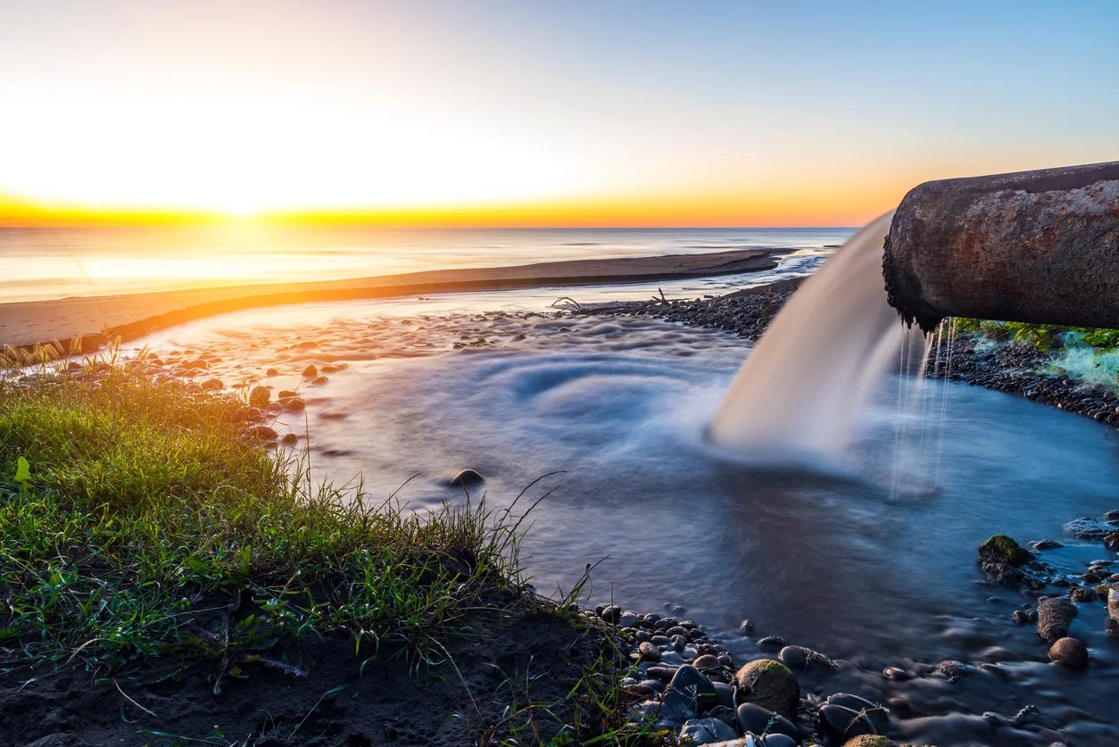 Sunset over an ocean beach with a pipe releasing water into a small stream, surrounded by rocks and grass.