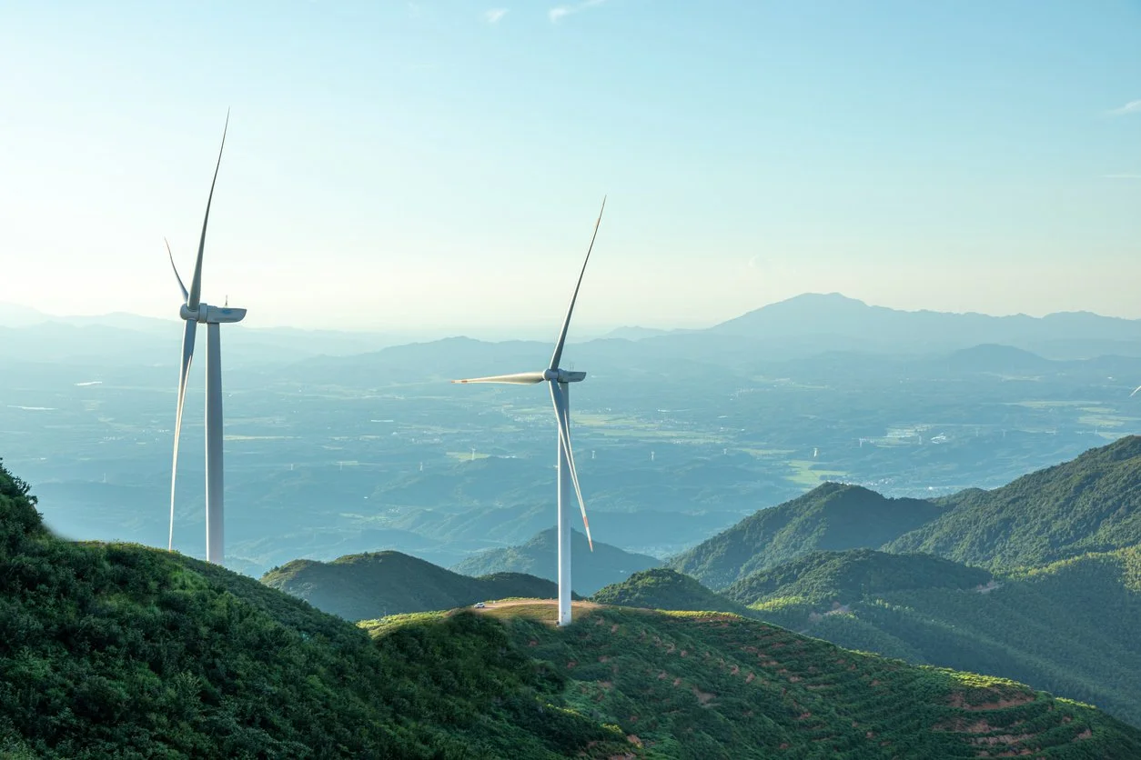Wind turbines on a lush green hillside overlooking a valley and distant mountains under a clear sky.