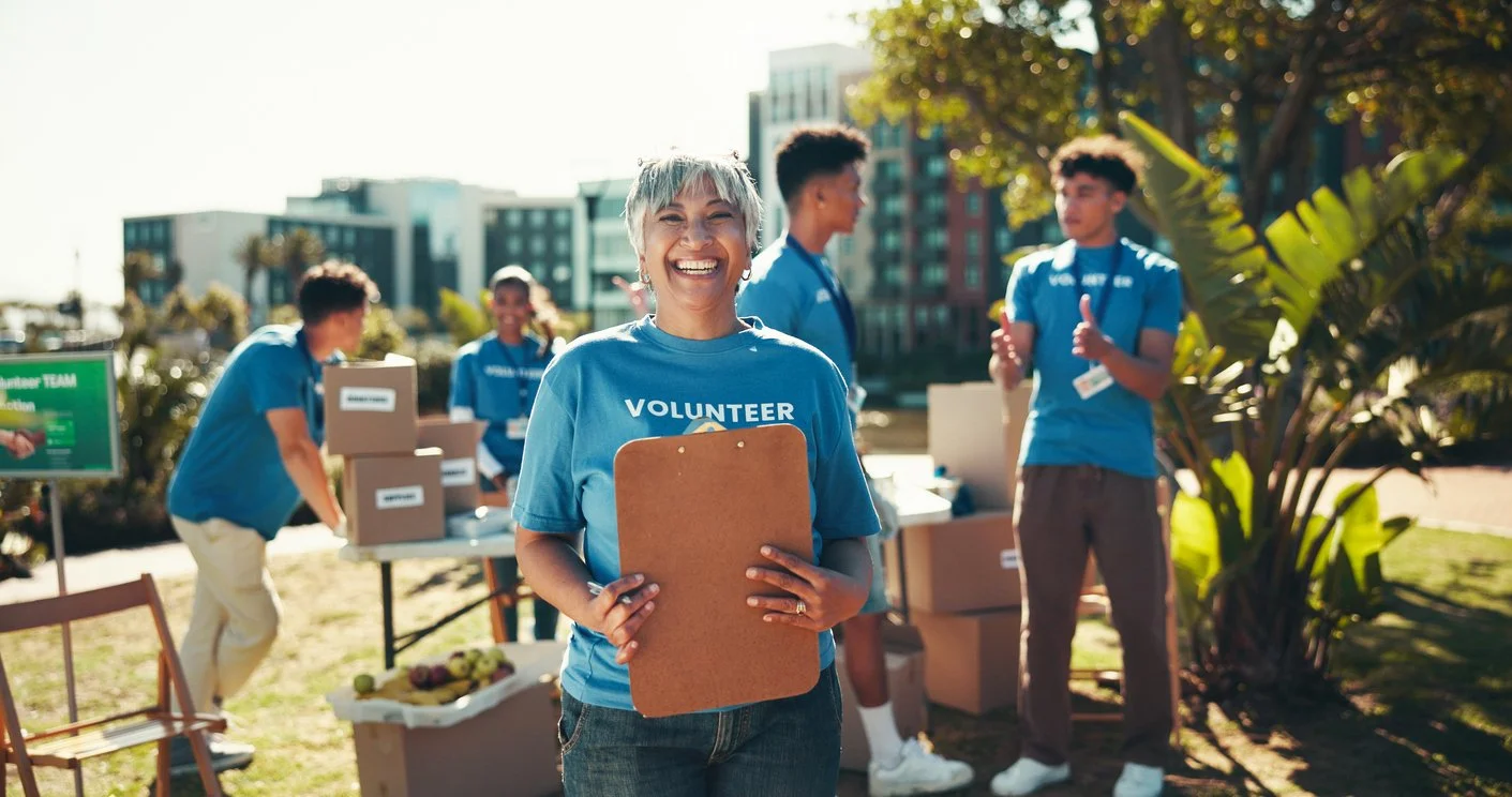 A smiling woman wearing a blue volunteer shirt holding a clipboard at an outdoor community event with other volunteers in the background.