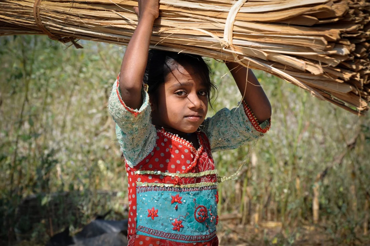 A young girl carrying a bundle of large, dry leaves on her head outdoors in a natural setting.