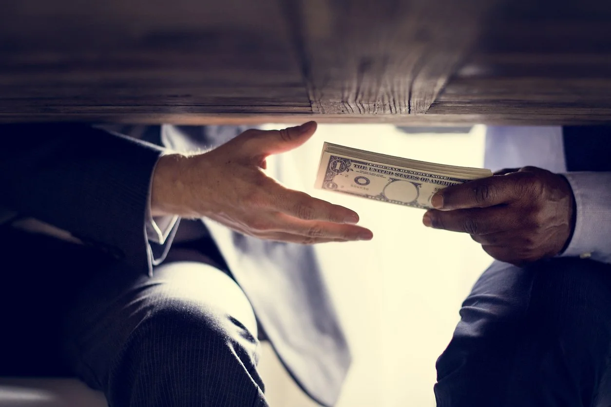 Two people exchanging a stack of hundred-dollar bills under a wooden table.