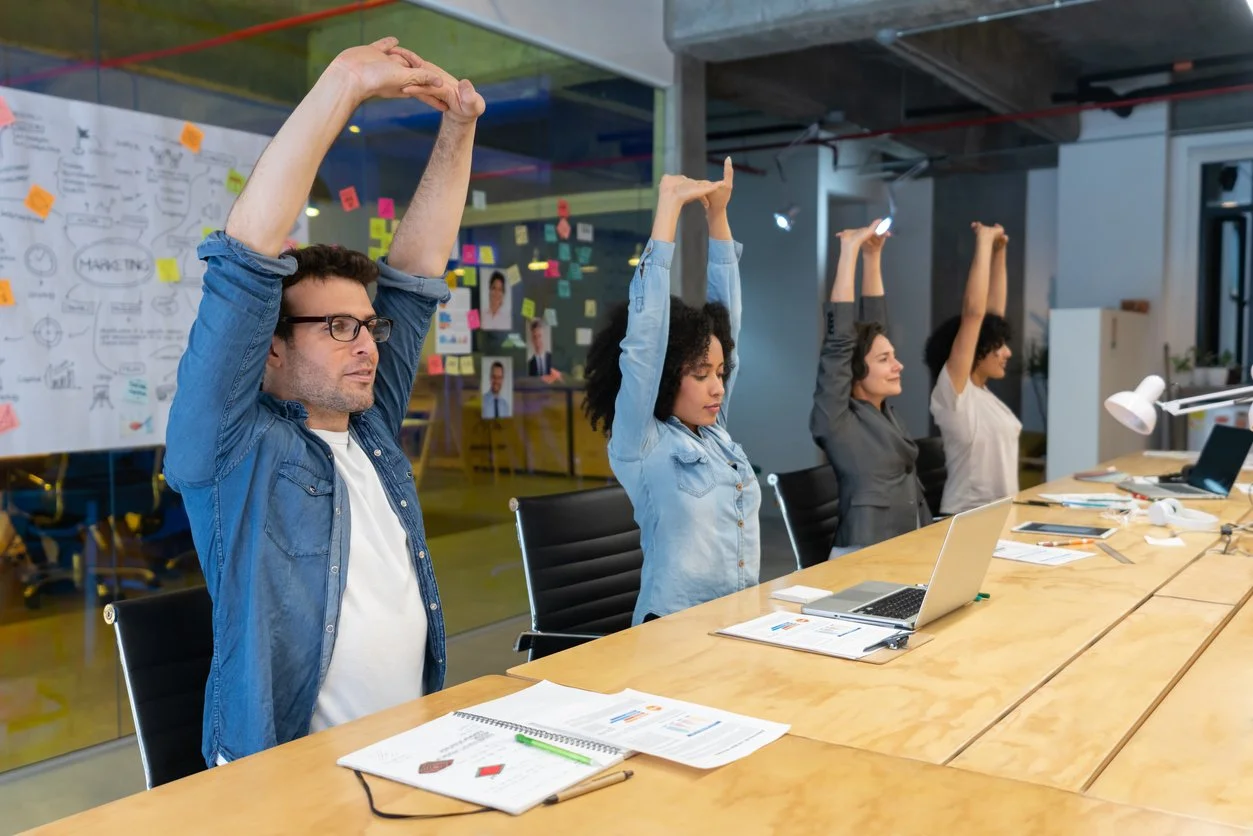Four people sitting at a conference table with laptops and documents, participating in a stretching exercise in a modern office, with a whiteboard and colorful sticky notes in the background.
