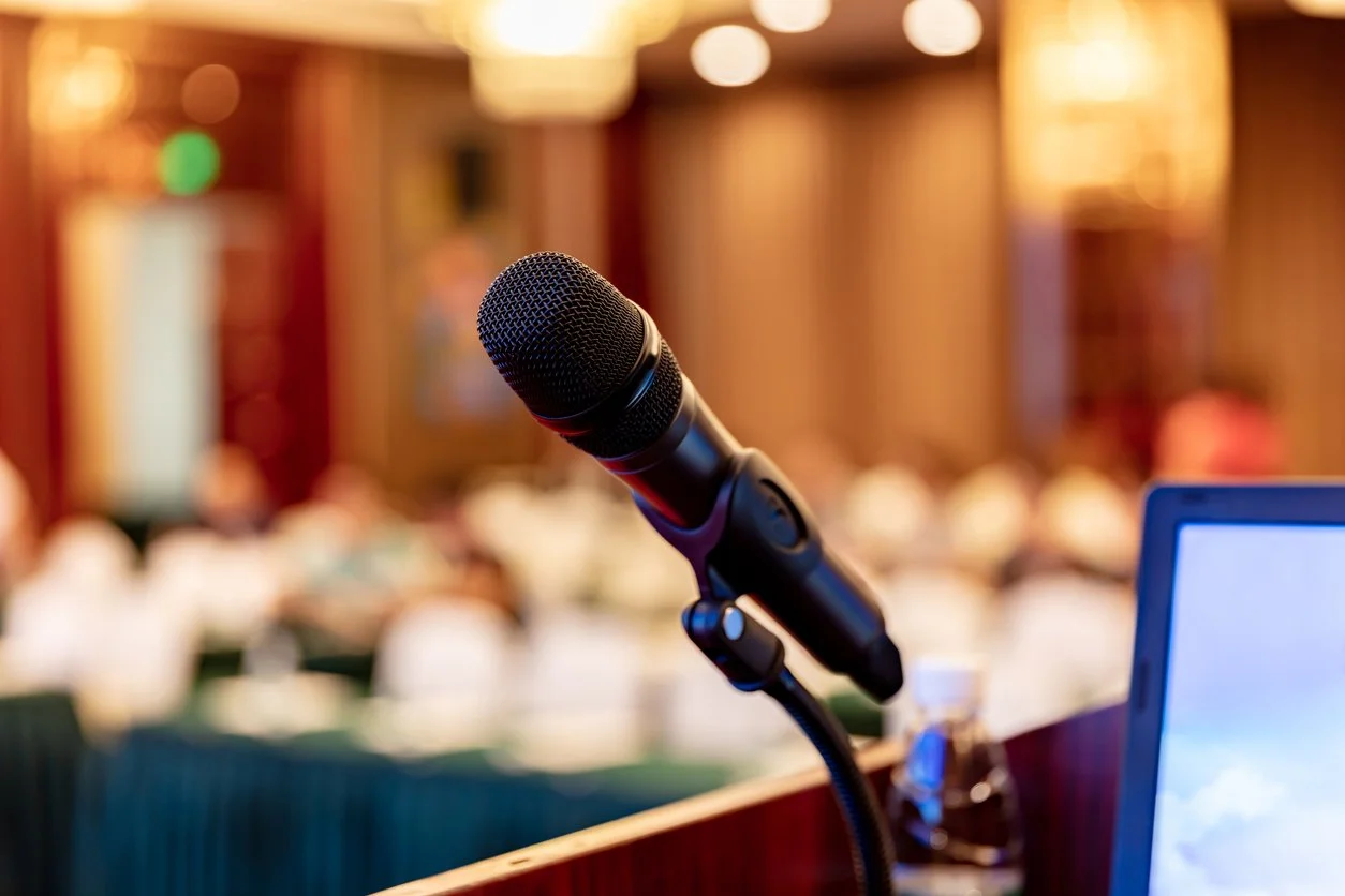 Microphone on stand in a conference room or auditorium, with blurred audience members and warm lighting in the background.