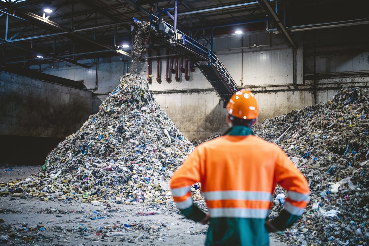 A worker in an orange safety uniform and hard hat standing with hands on hips, overseeing a large pile of mixed recyclable waste in an industrial recycling facility. A conveyor belt is dropping waste onto the pile."
