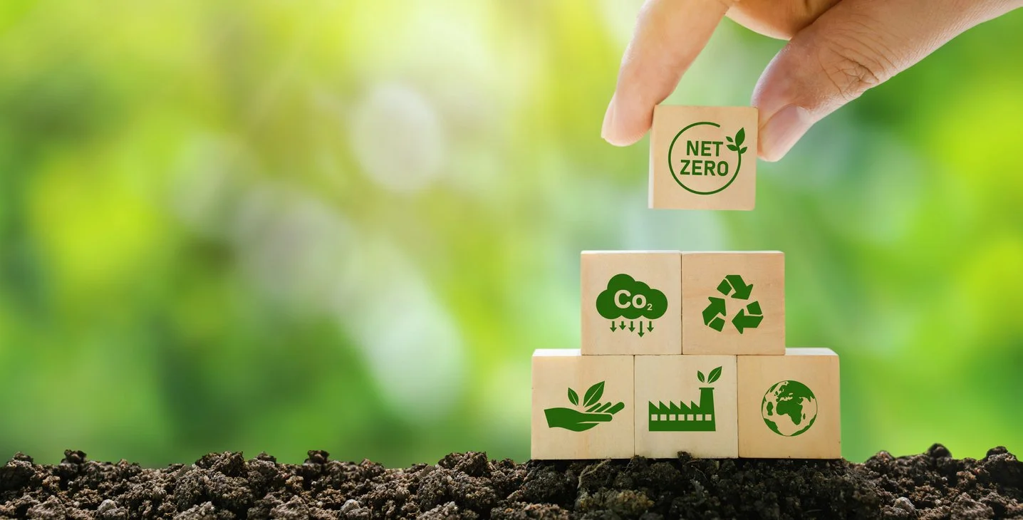 Hand placing a block with 'NET ZERO' and a leaf symbol on top of a structure made of wooden blocks, with environmental icons such as CO2 reduction, recycling, planting, and Earth on the blocks, against a blurred green background.
