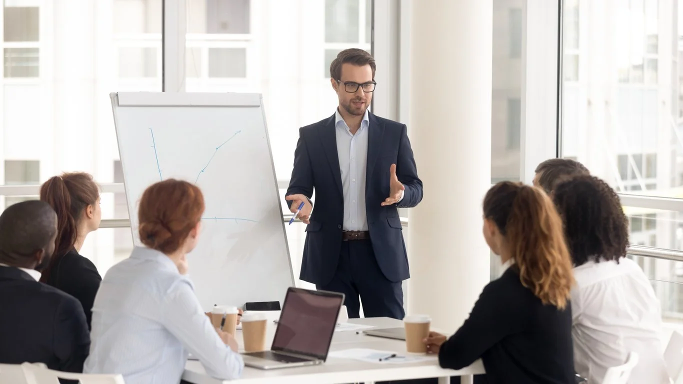 Businessman in suit giving a presentation to a diverse group of professionals in a modern office conference room.