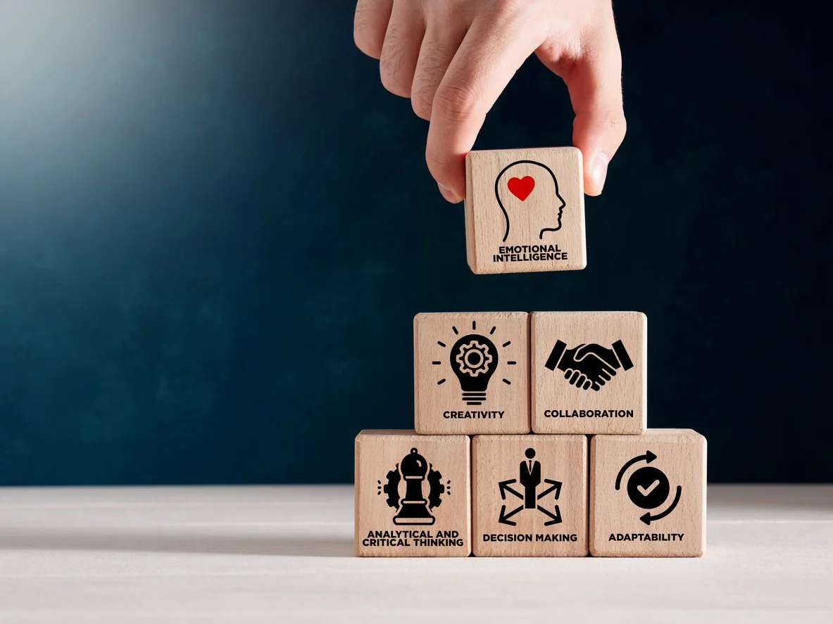 Hand placing a wooden block labeled 'Emotional Intelligence' above a pyramid of other wooden blocks labeled 'Creativity,' 'Collaboration,' 'Analytical and Critical Thinking,' 'Decision Making,' and 'Adaptability.'