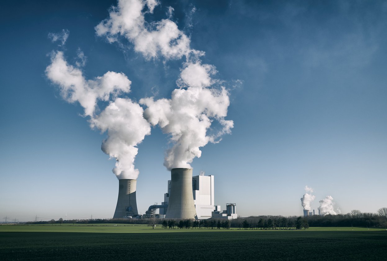 A power plant with cooling towers emitting steam, situated in a flat, green landscape under a blue sky with scattered clouds.