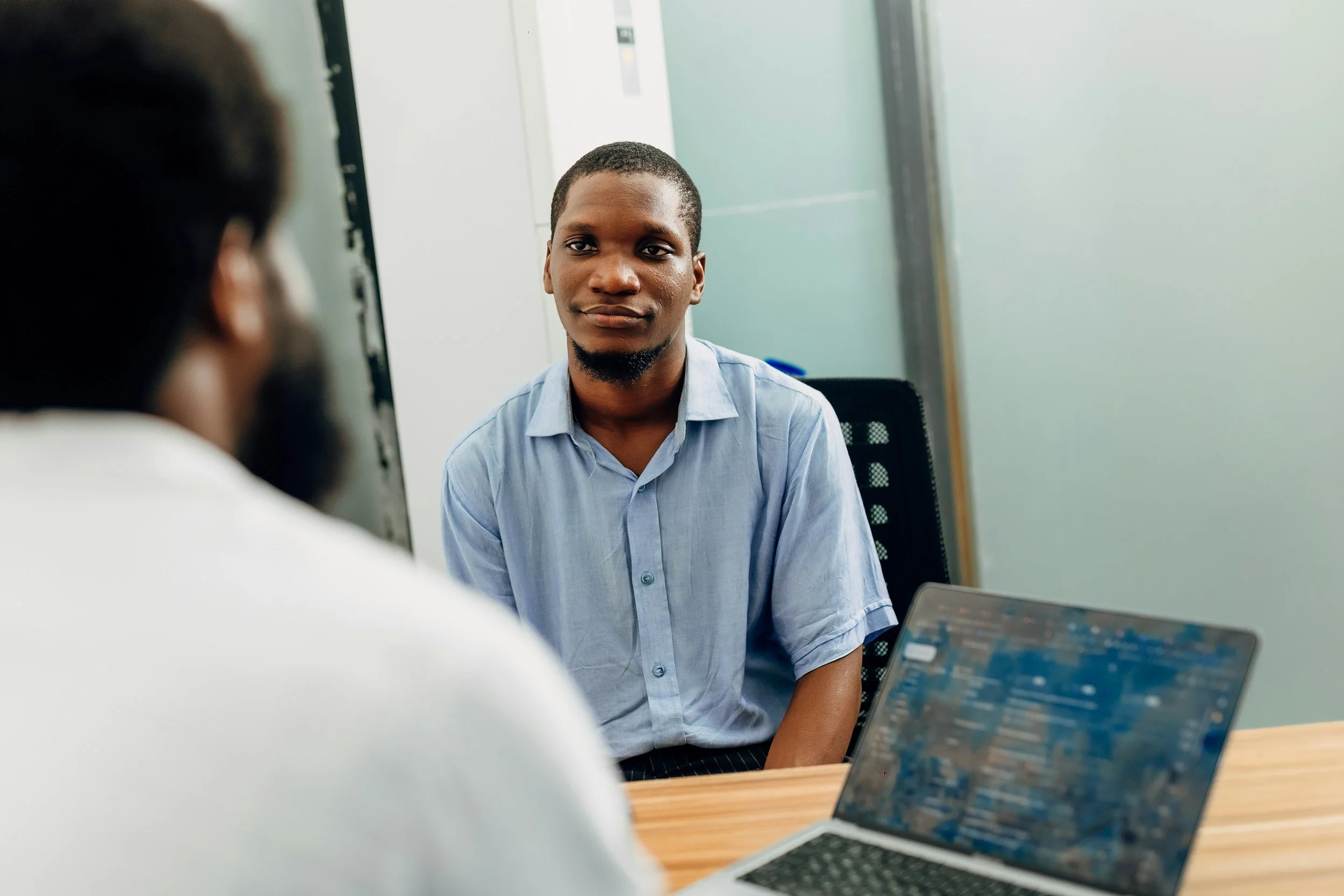 Two men having a conversation in an office setting, one facing the camera with a laptop open on the desk.