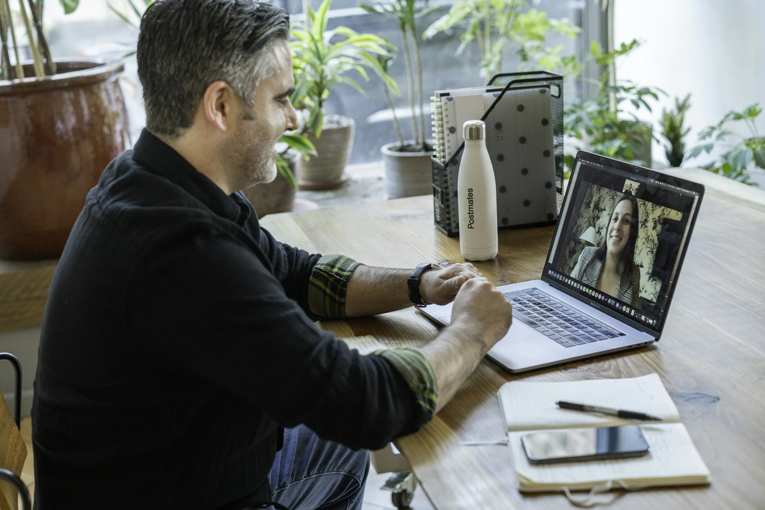 A man is having a video call on his laptop with a woman who is smiling. The man is sitting at a wooden table with a notebook, a pen, and a smartphone in front of him. The setting appears to be a bright, plant-filled room.
