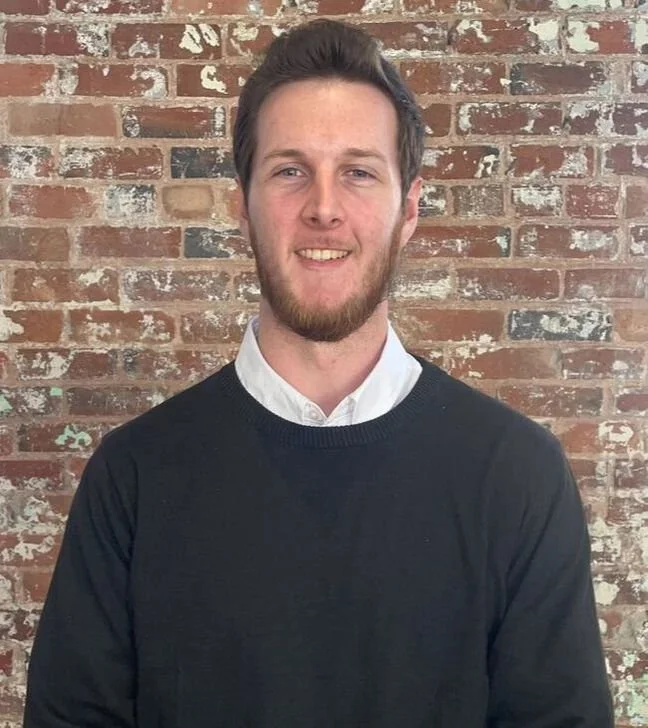 A young man with short brown hair and a beard, wearing a white collared shirt under a black sweater, standing in front of a brick wall.