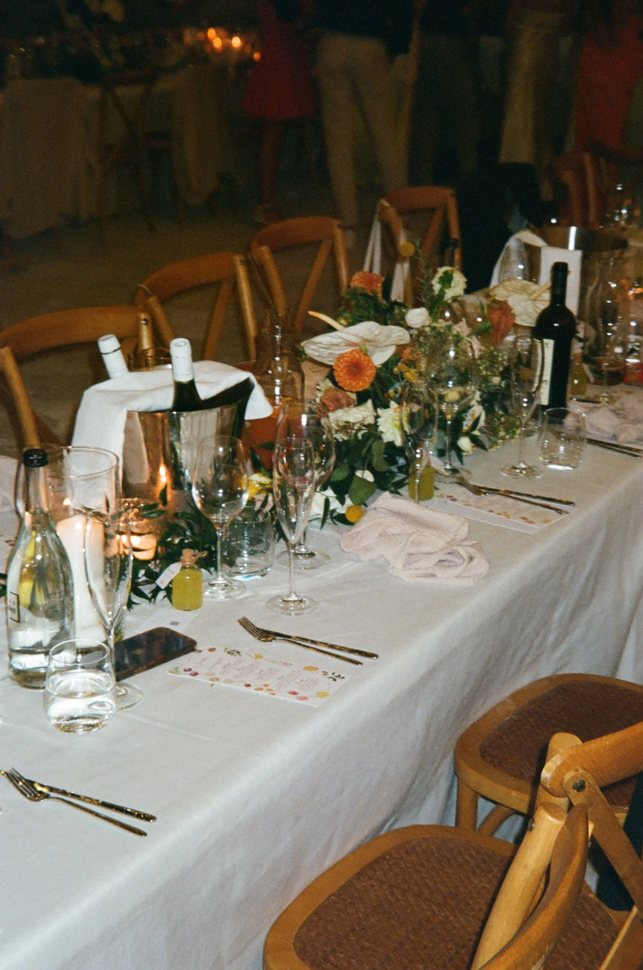 A decorated banquet table set for a formal event with wine bottles, glasses, floral arrangements, and candles, in a dimly lit room.