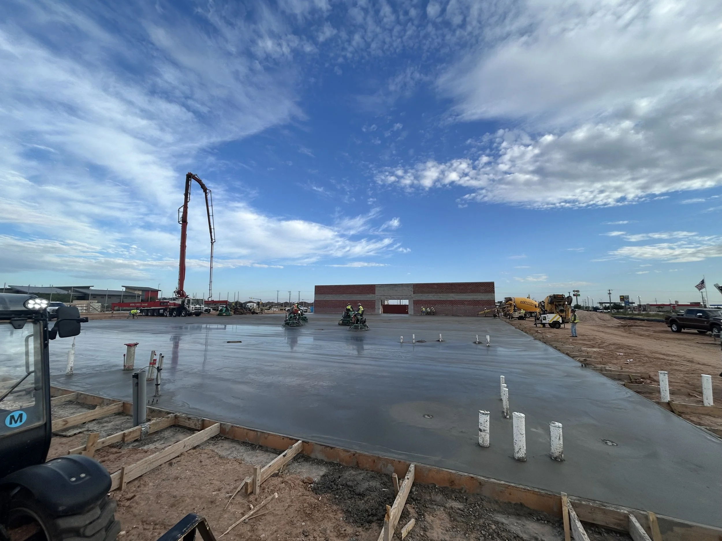 Construction site with freshly poured concrete and workers. Equipment and materials visible, including a concrete pump truck and construction machinery under a partly cloudy sky.