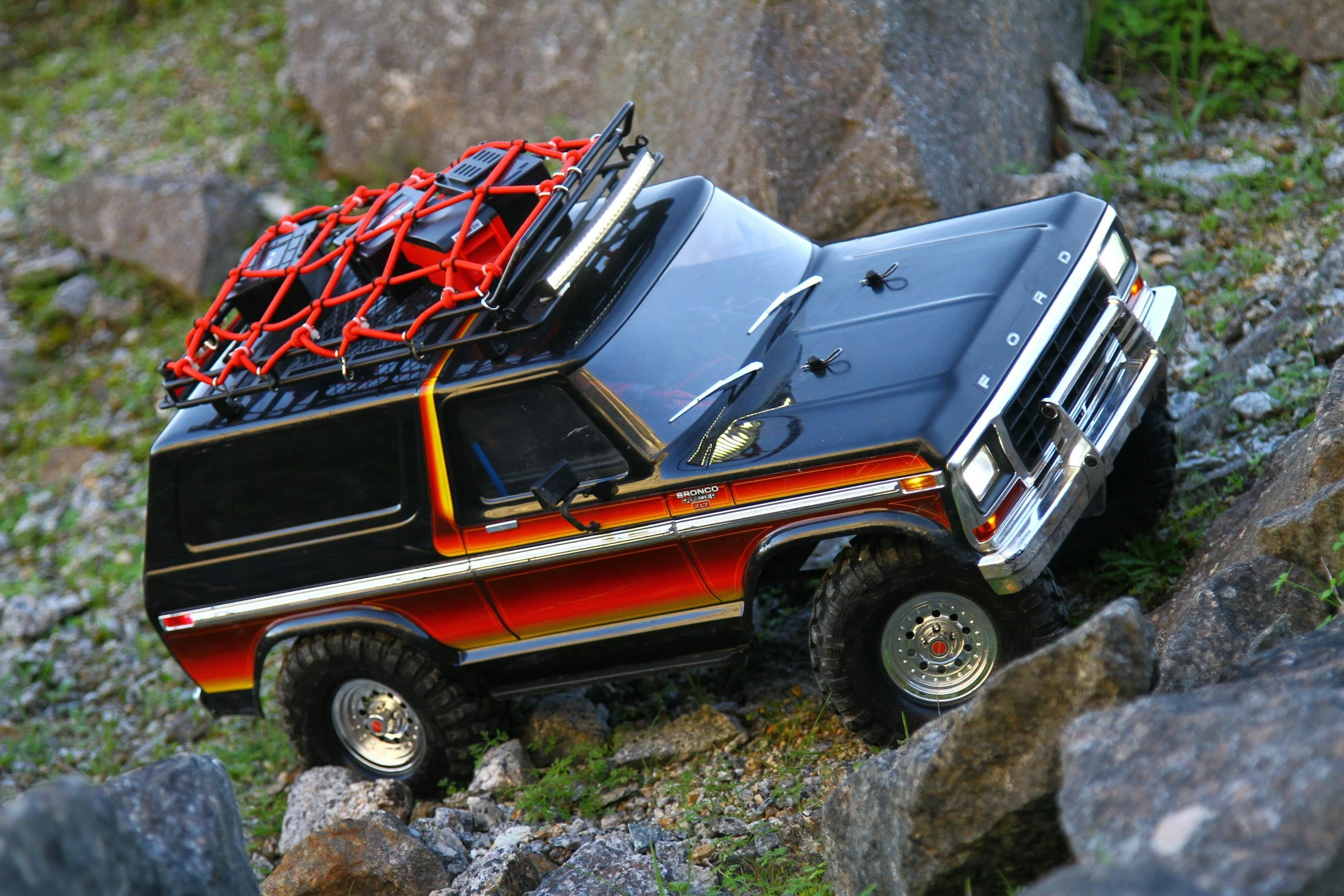 A black and red off-road toy vehicle ascending a rocky terrain with a top-mounted red cargo net and a small antenna on the hood.