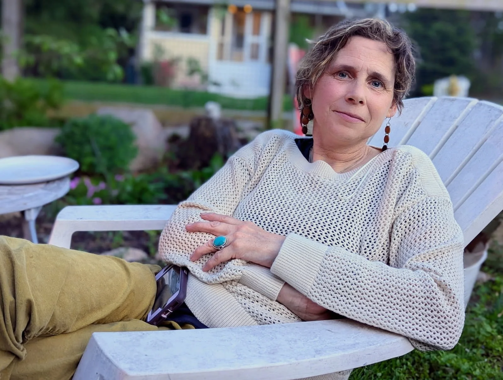 A woman sitting outdoors in a white Adirondack chair, wearing a cream-colored knit sweater, earrings, and a turquoise ring. She has short, wavy hair and is resting her arms on a white table.