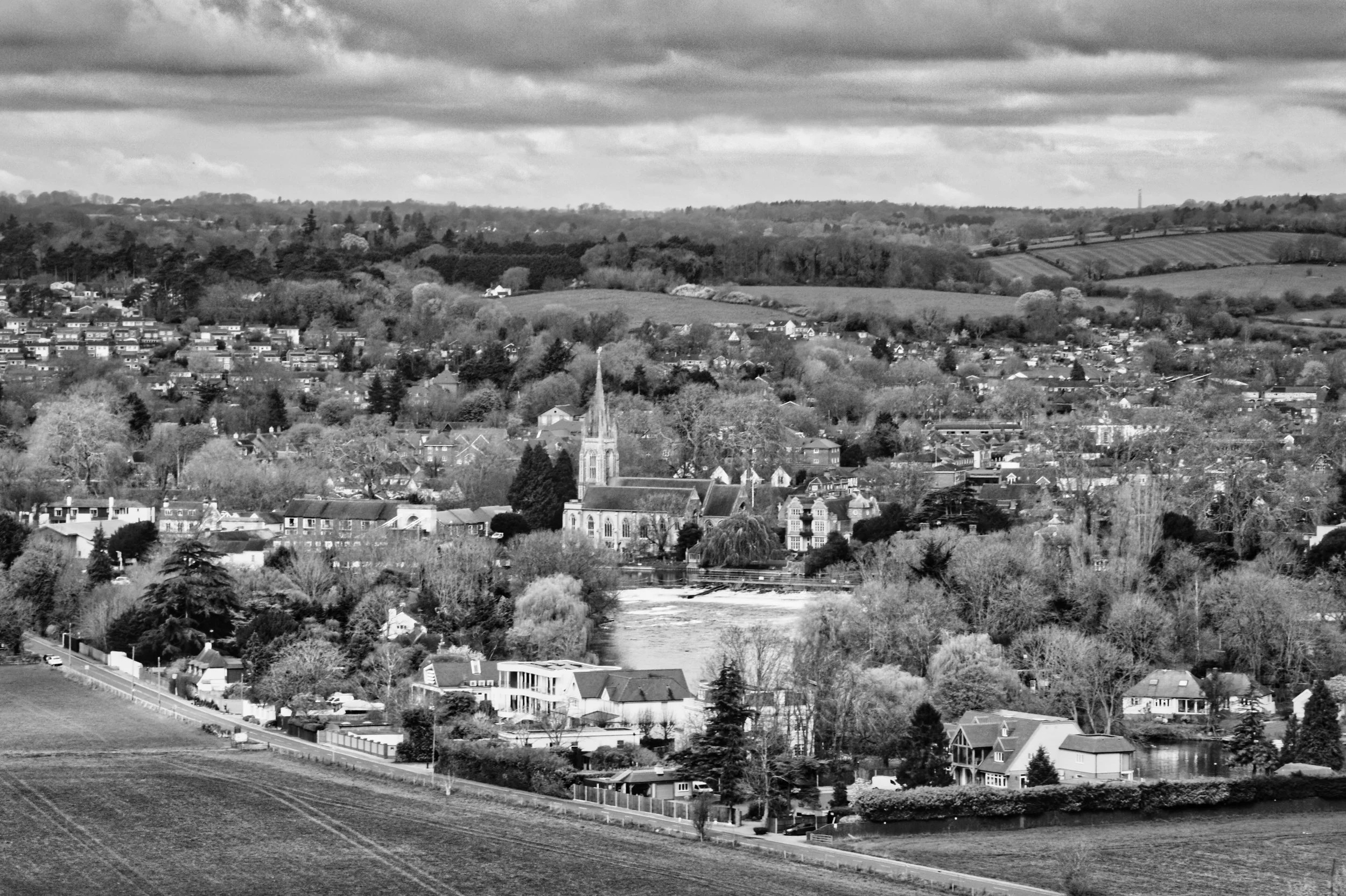 A black and white aerial view over Marlow, Buckinghamshire, from Quarry Wood, showing All Saints Church spire rising above the town with the River Thames weir visible and the Chiltern Hills in the background.