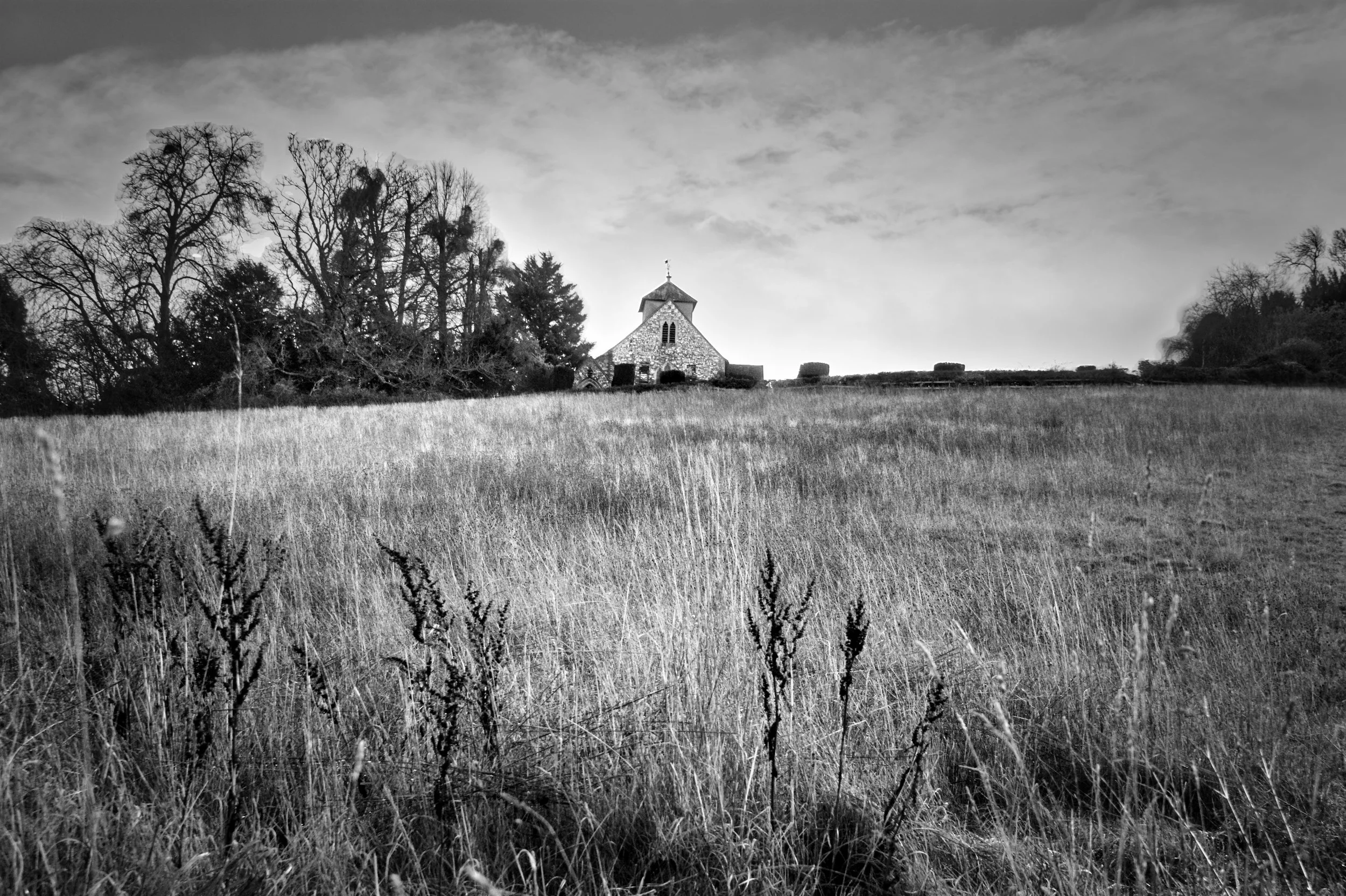 Black and white photograph of St Nicholas Church on a hillside at Hedsor, viewed across long winter grass under a dramatic sky.