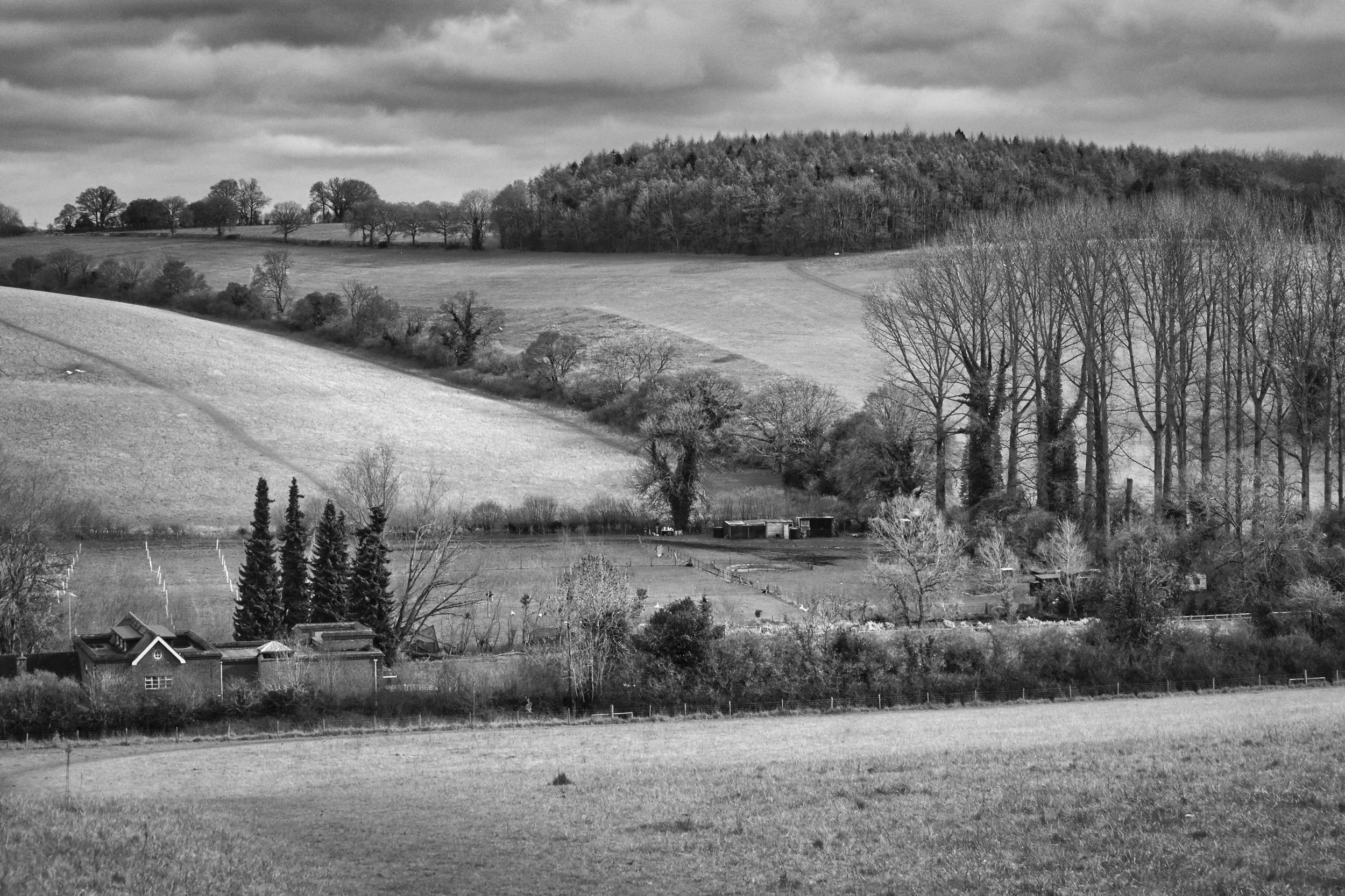 Rolling valley farmland behind Chorleywood House Estate looking towards Blunts Wood, farm buildings and bare trees in the valley floor, dramatic cloudy sky, Chess Valley, Buckinghamshire, black and white