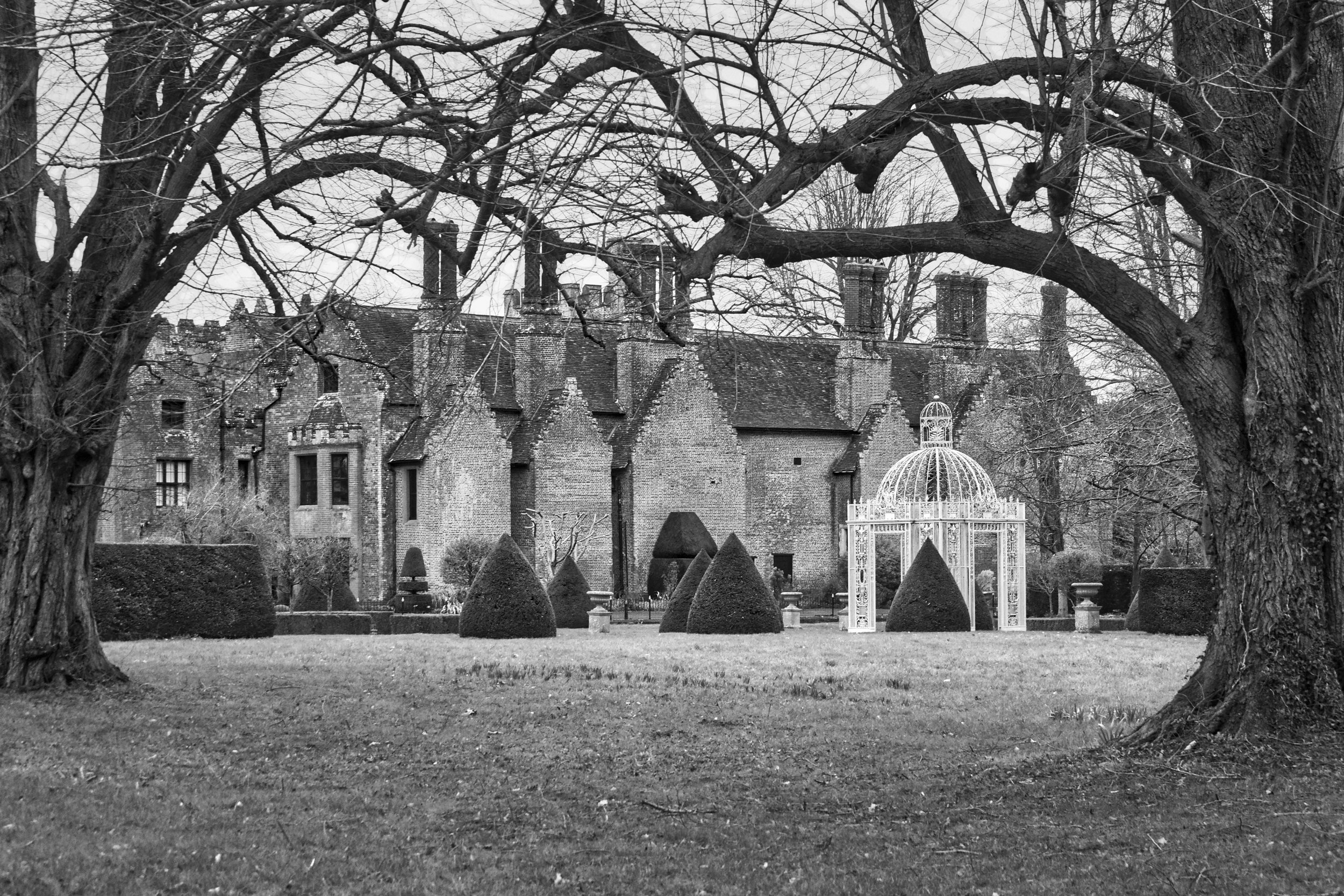 Chenies Manor House viewed through bare trees, Tudor brick facade with topiary gardens and ornate white gazebo in the foreground, Buckinghamshire, black and white