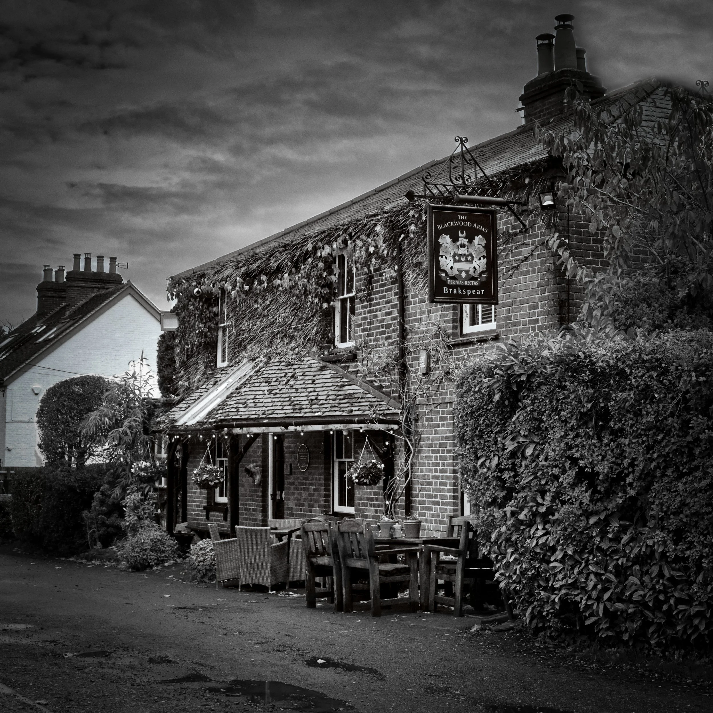 Black and white photograph of an ivy-covered brick village pub, The Blackwood Arms, with outdoor tables and chairs beneath a cloudy sky in the Chiltern Hills.