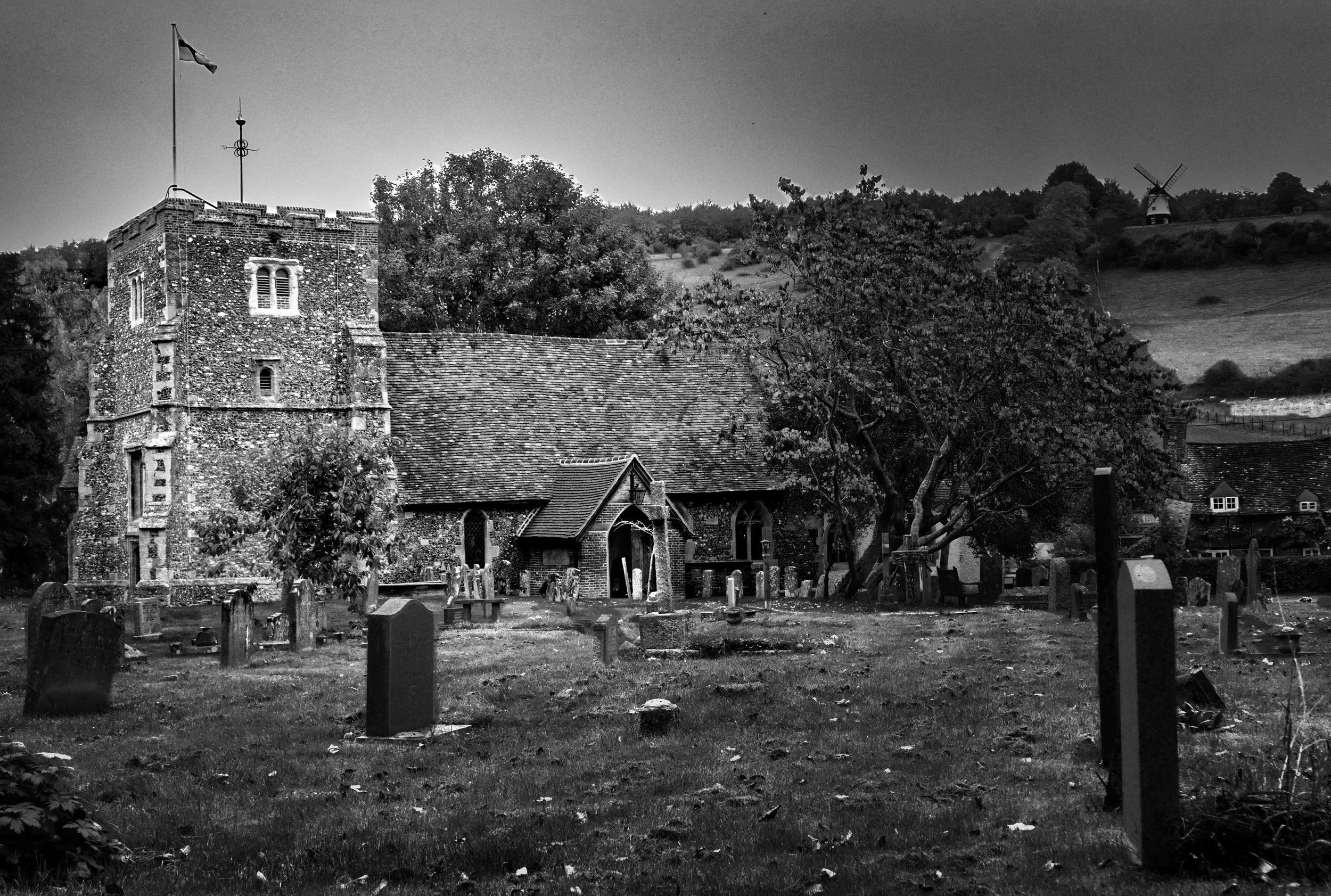 Black and white photograph of St Mary’s Church in Turville, with gravestones in the foreground and Cobstone Windmill visible on the hillside behind, known from The Vicar of Dibley and Chitty Chitty Bang Bang.