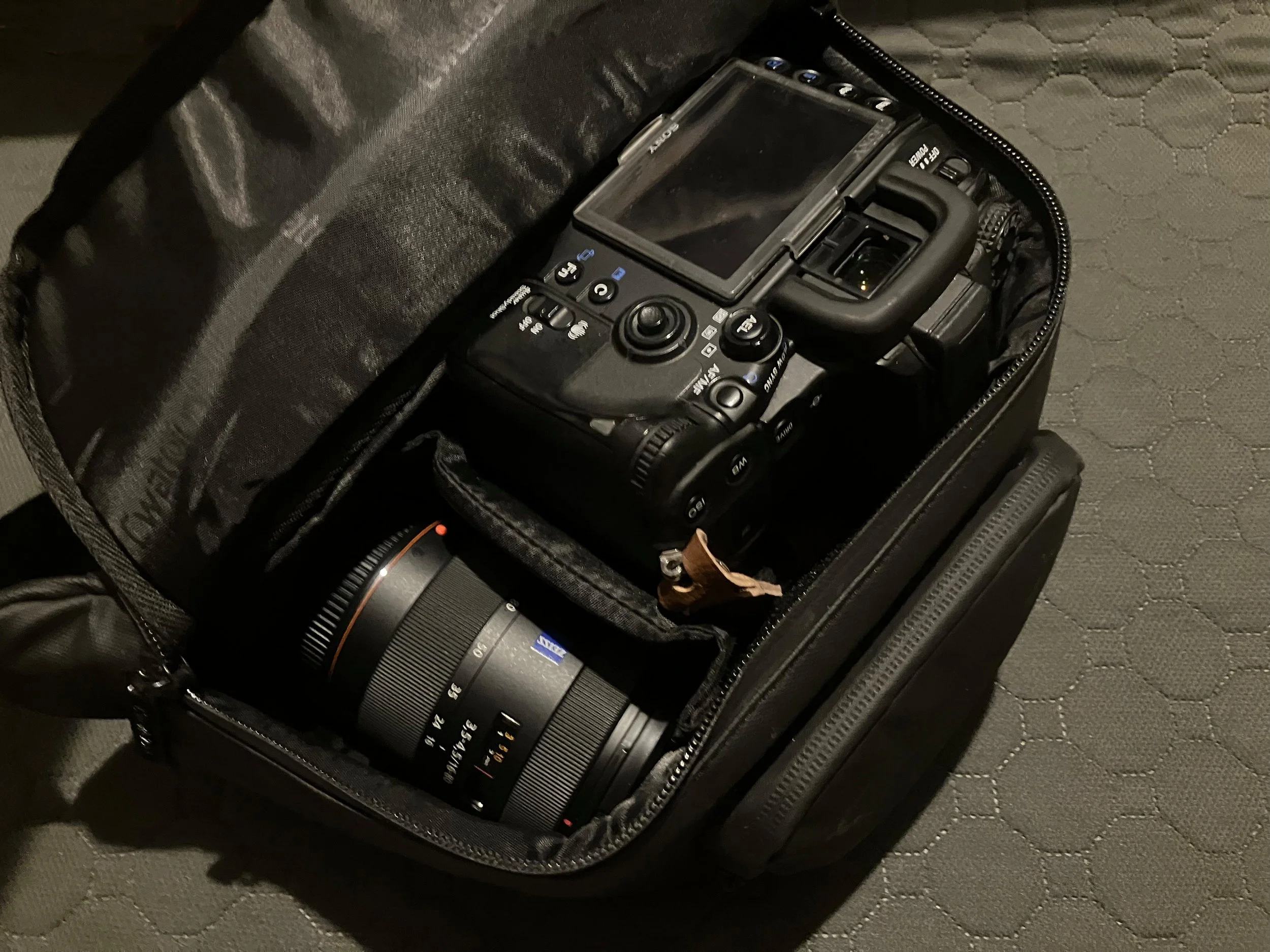 Sony A700 DSLR camera sitting inside a black hip pack, with a Carl Zeiss lens stored alongside it, viewed from above on a textured mat.