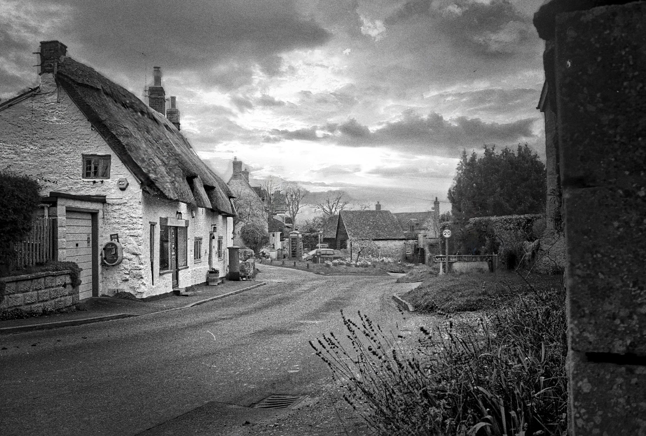 Black and white photograph of Chiltern village street with stone cottages, thatched roofs and a curved road leading downhill. By Mark Weekes