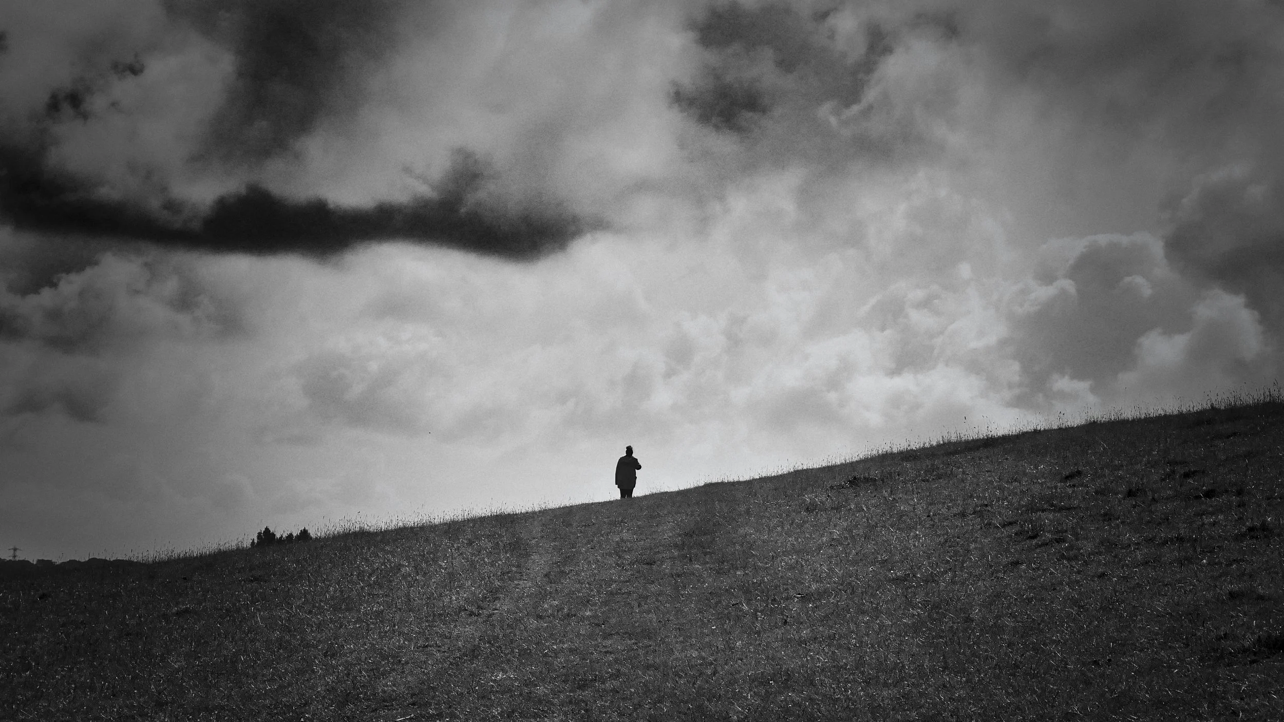 Black and white photograph of a solitary figure silhouetted on a hillside under a dramatic stormy sky, South Downs, England