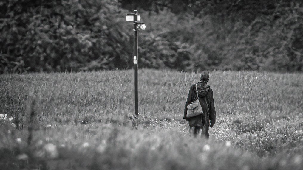 Black and white image of a woman standing alone in the middle of a field, staring at a signpost, evoking reflection and curiosity.