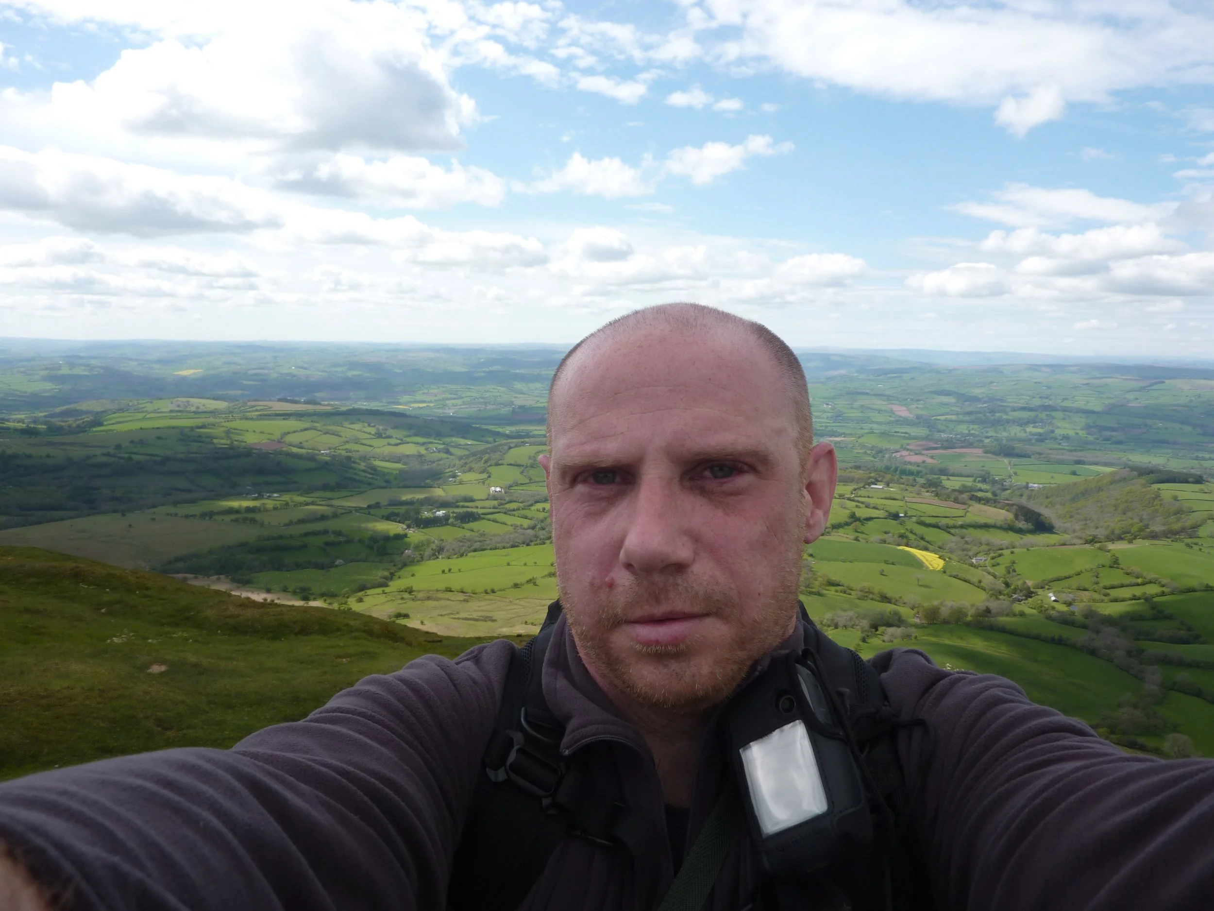 A man taking a selfie on a hilltop with a vast green landscape and cloudy sky in the background.