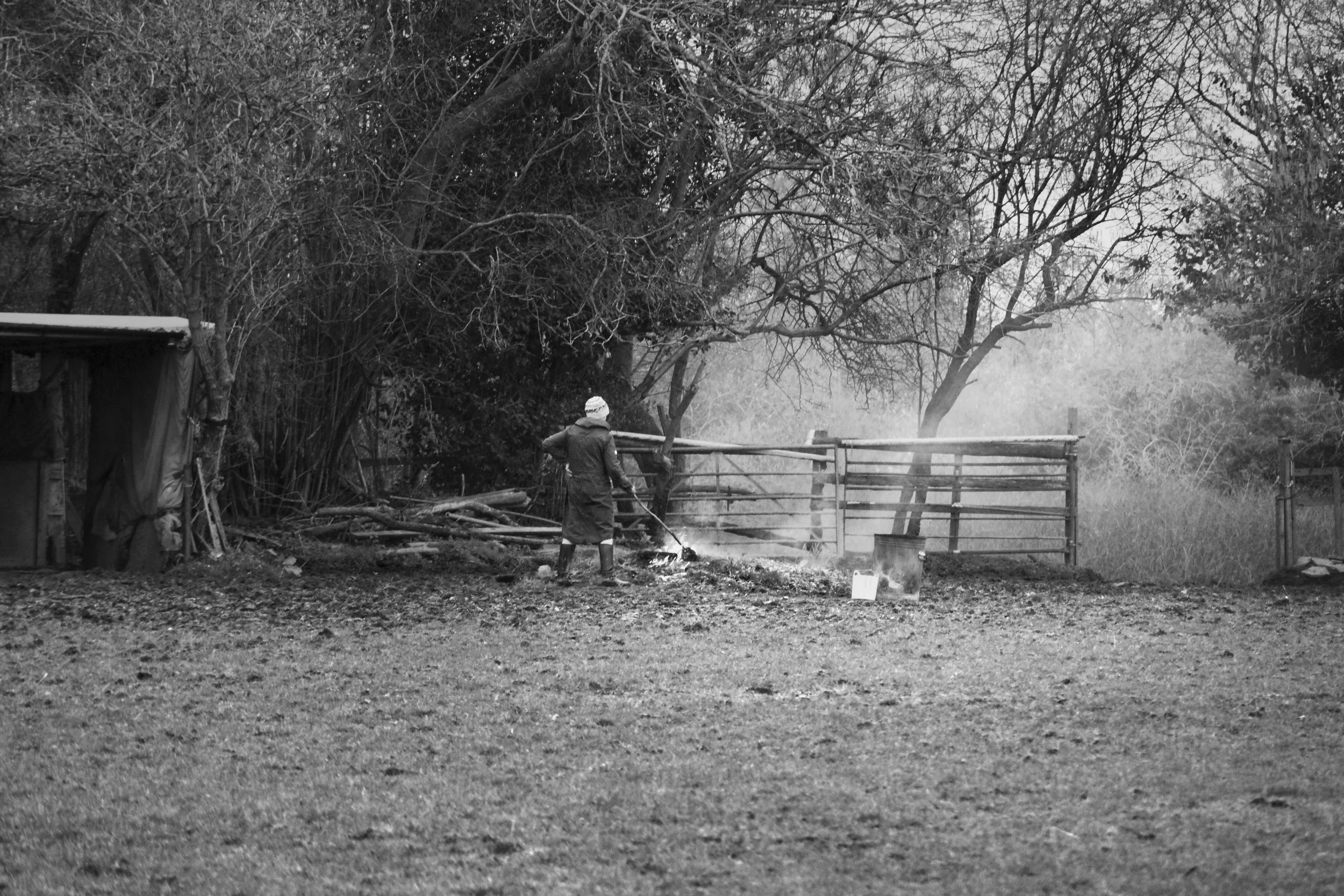 Farm worker at a wooden gate in a rural farmyard, smoke rising from a fire, trees behind, black and white