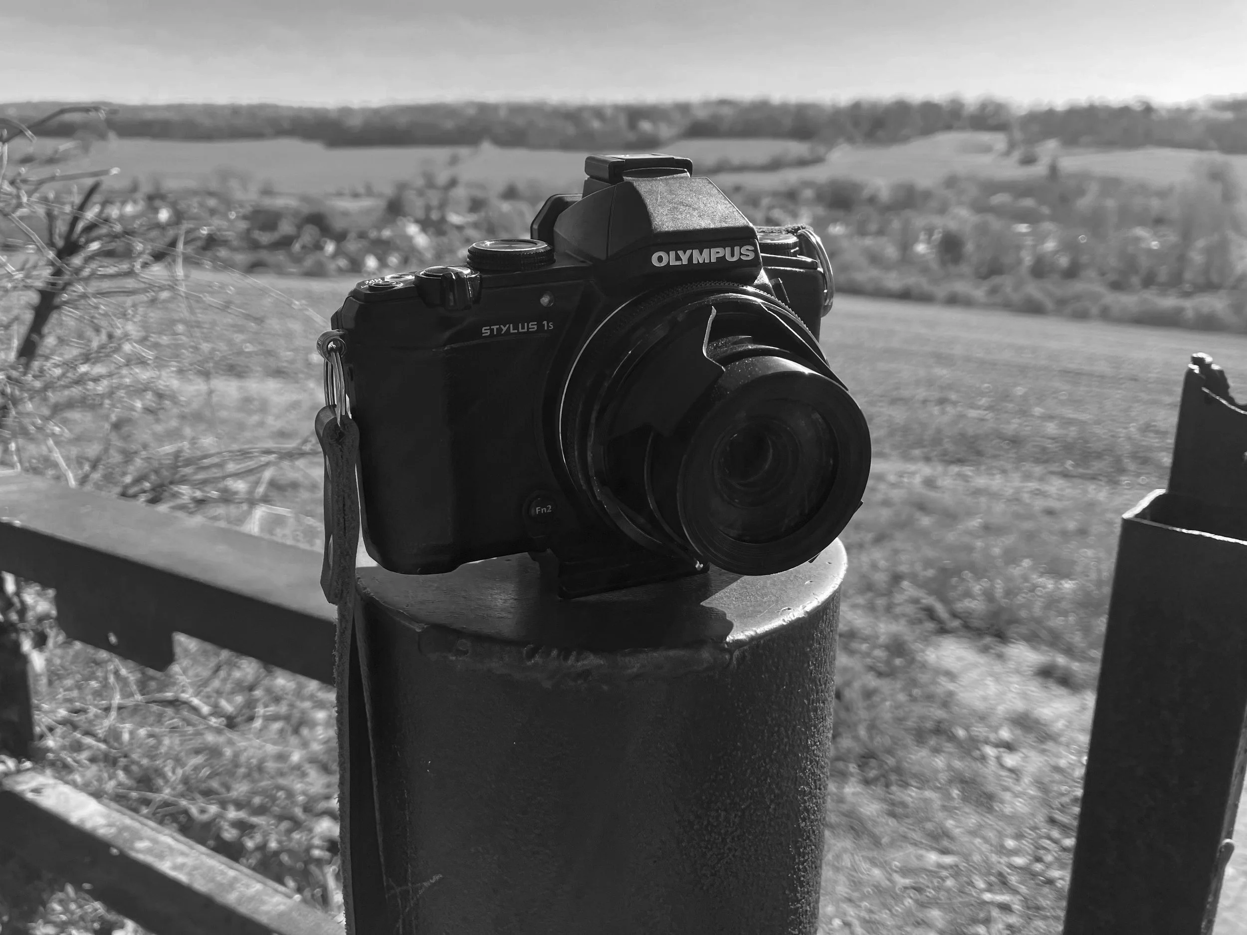 Black and white image of an Olympus Stylus 1s sat on top of a post in the chilterns countryside
