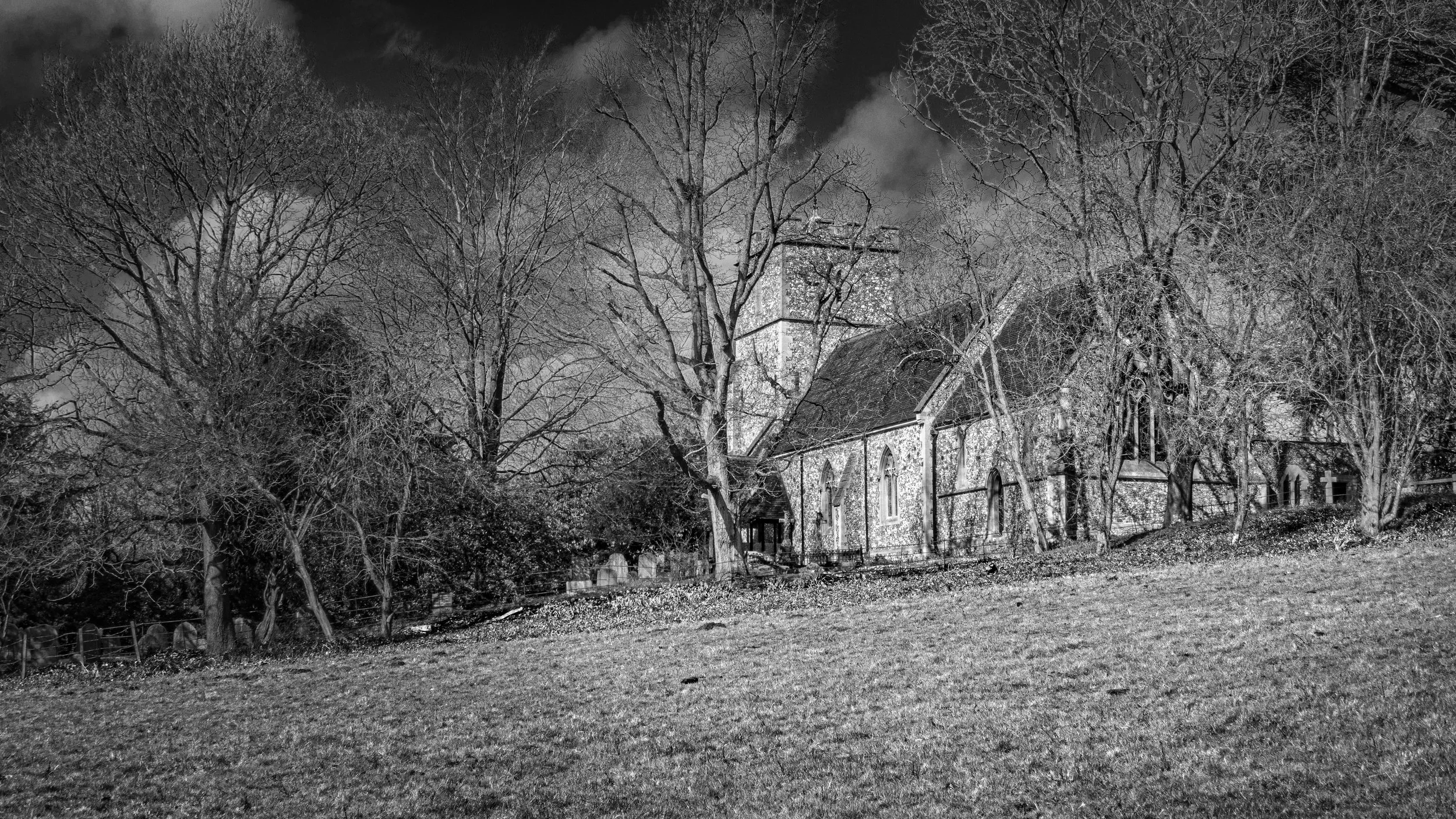 Black and white photograph of Church Wood in Buckinghamshire, showing a flint village church partly hidden among bare winter trees beneath a cloudy sky.