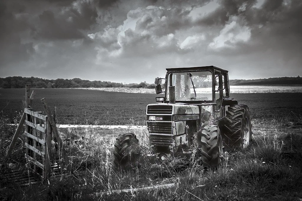 An abandoned rusting tractor sits overgrown with weeds beside a broken wooden gate at Little Marlow, Buckinghamshire, dramatic storm clouds building over open farmland behind it.