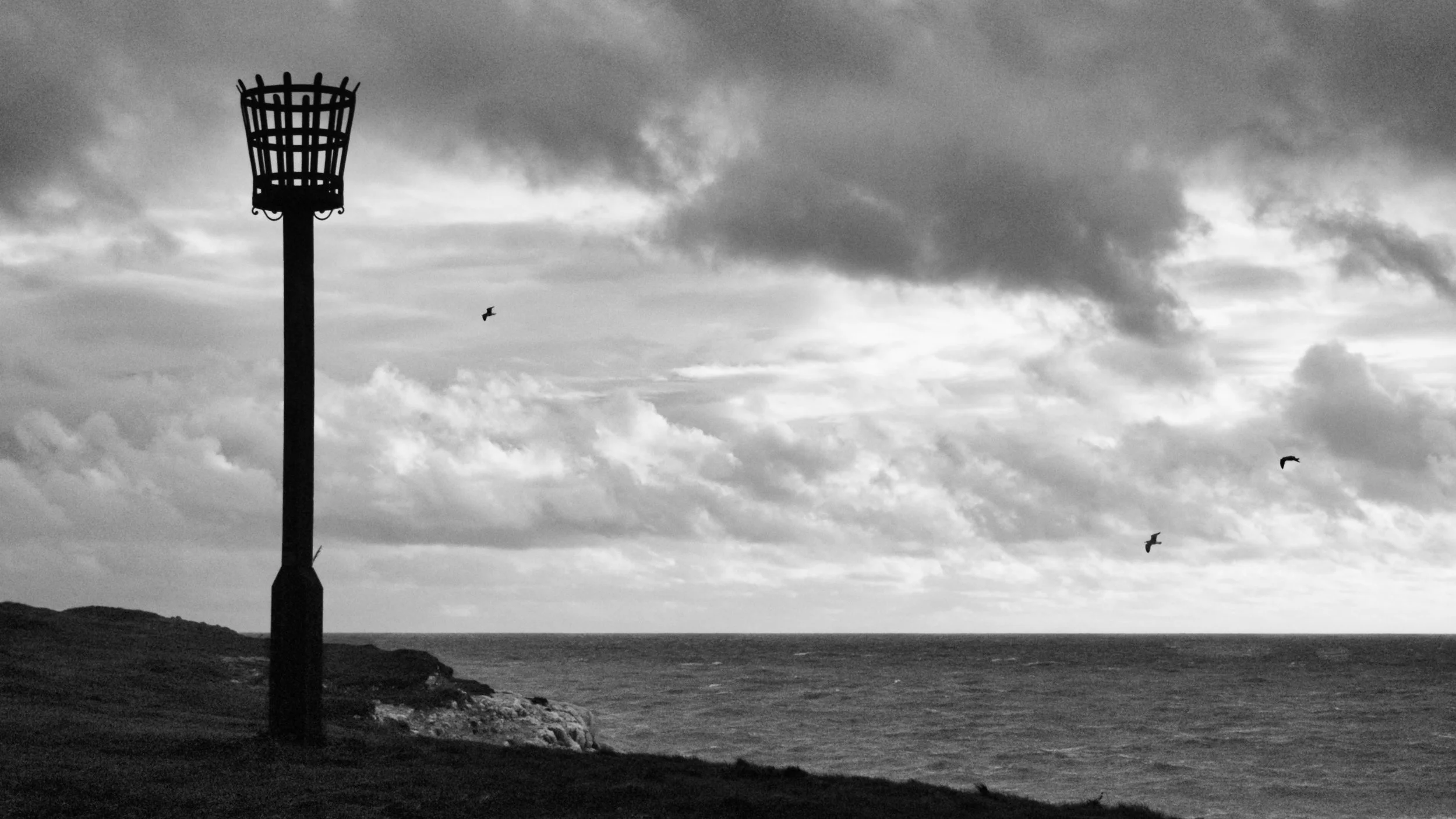 Black and white photograph of a coastal beacon overlooking the sea at Seaford, with birds flying under a cloudy sky.