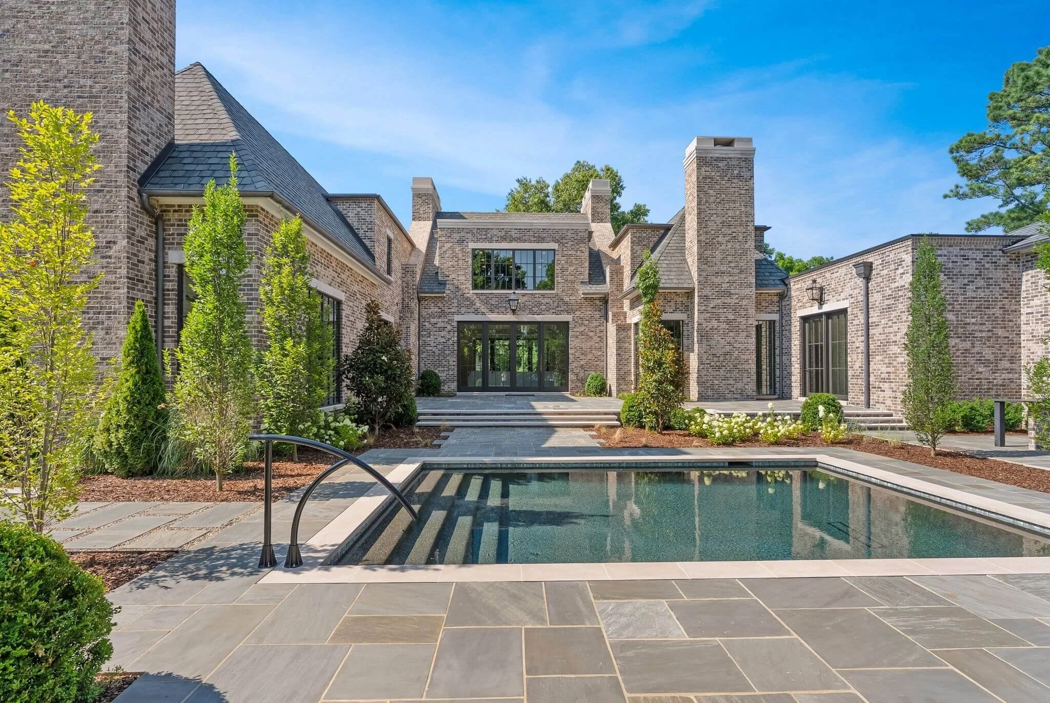 Front view of a large brick house with a swimming pool in the foreground, surrounded by greenery and trees, under a clear blue sky.
