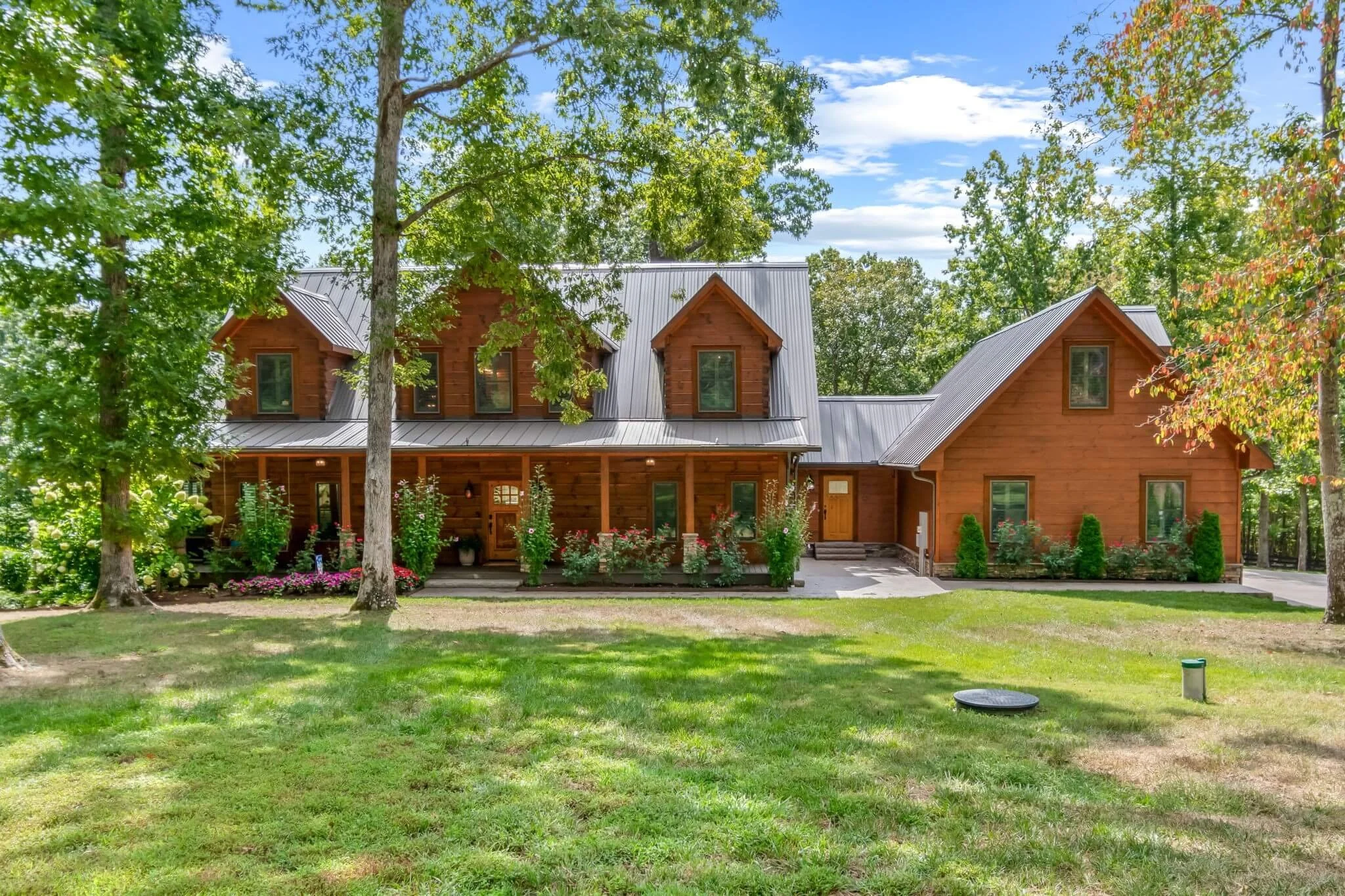 A large wooden house with a metal roof, surrounded by green trees and grass, with a front porch and a flower garden.