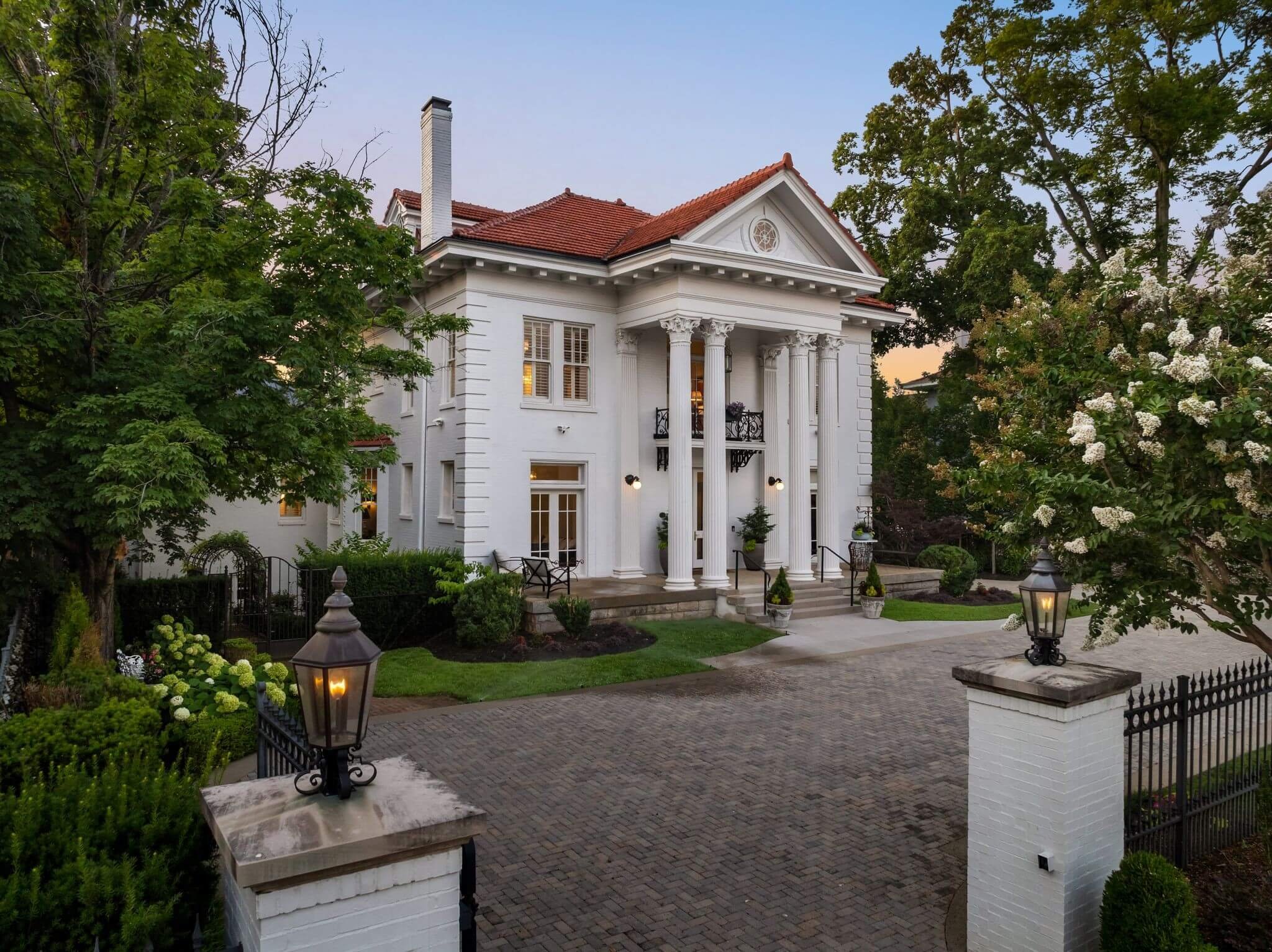 A large white mansion with tall columns at the entrance, surrounded by trees and lit lanterns in a landscaped yard.