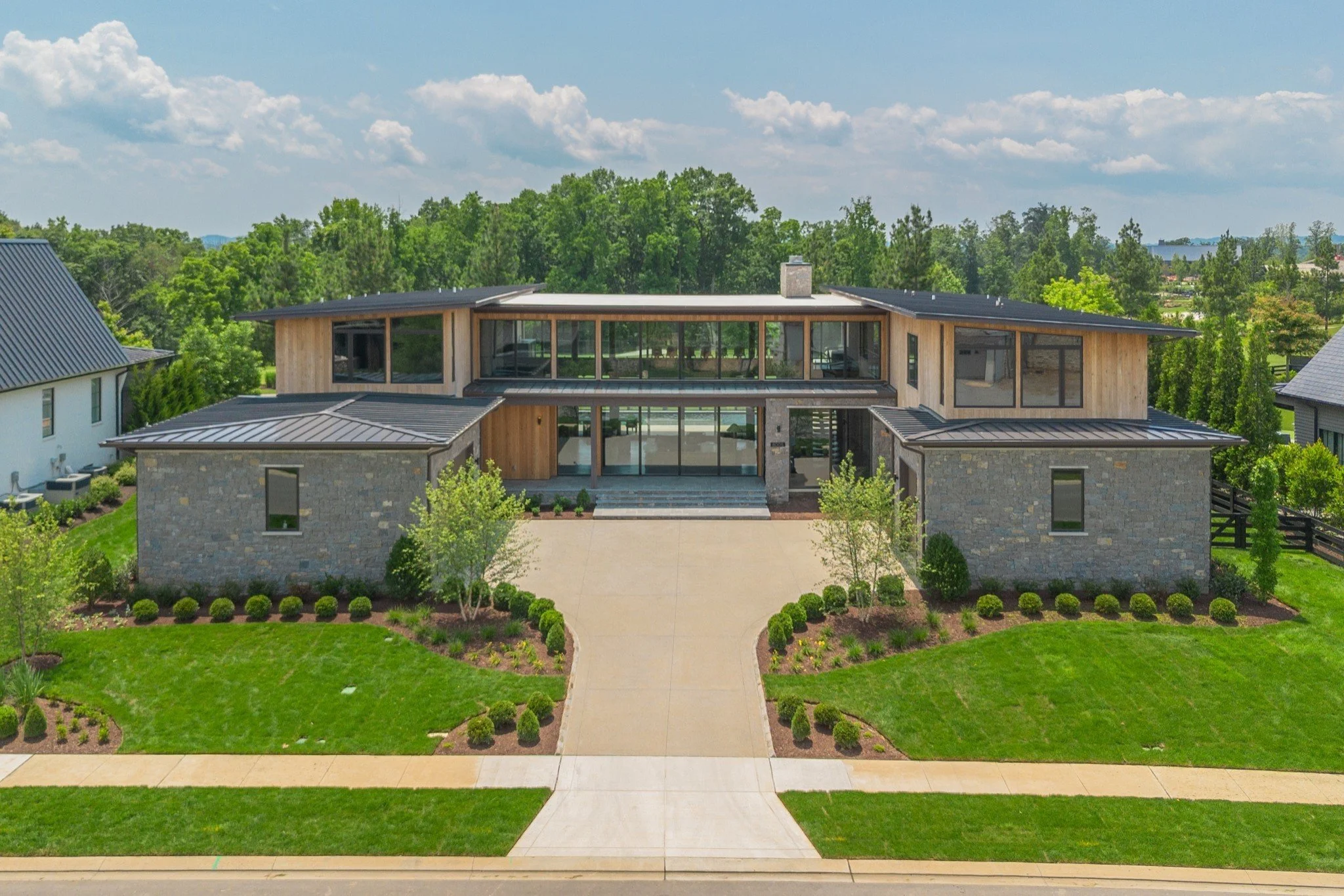 A modern two-story house with stone and wood exterior, large glass windows, a front driveway, and landscaped yard with bushes and small trees.