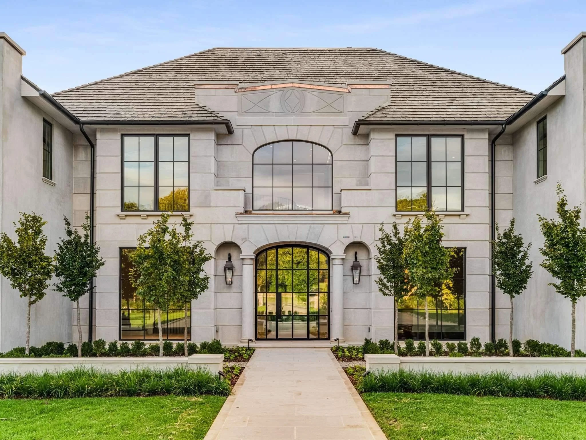 Front view of a modern two-story house with large windows, a stone pathway, and landscaped greenery including trees and shrubs.