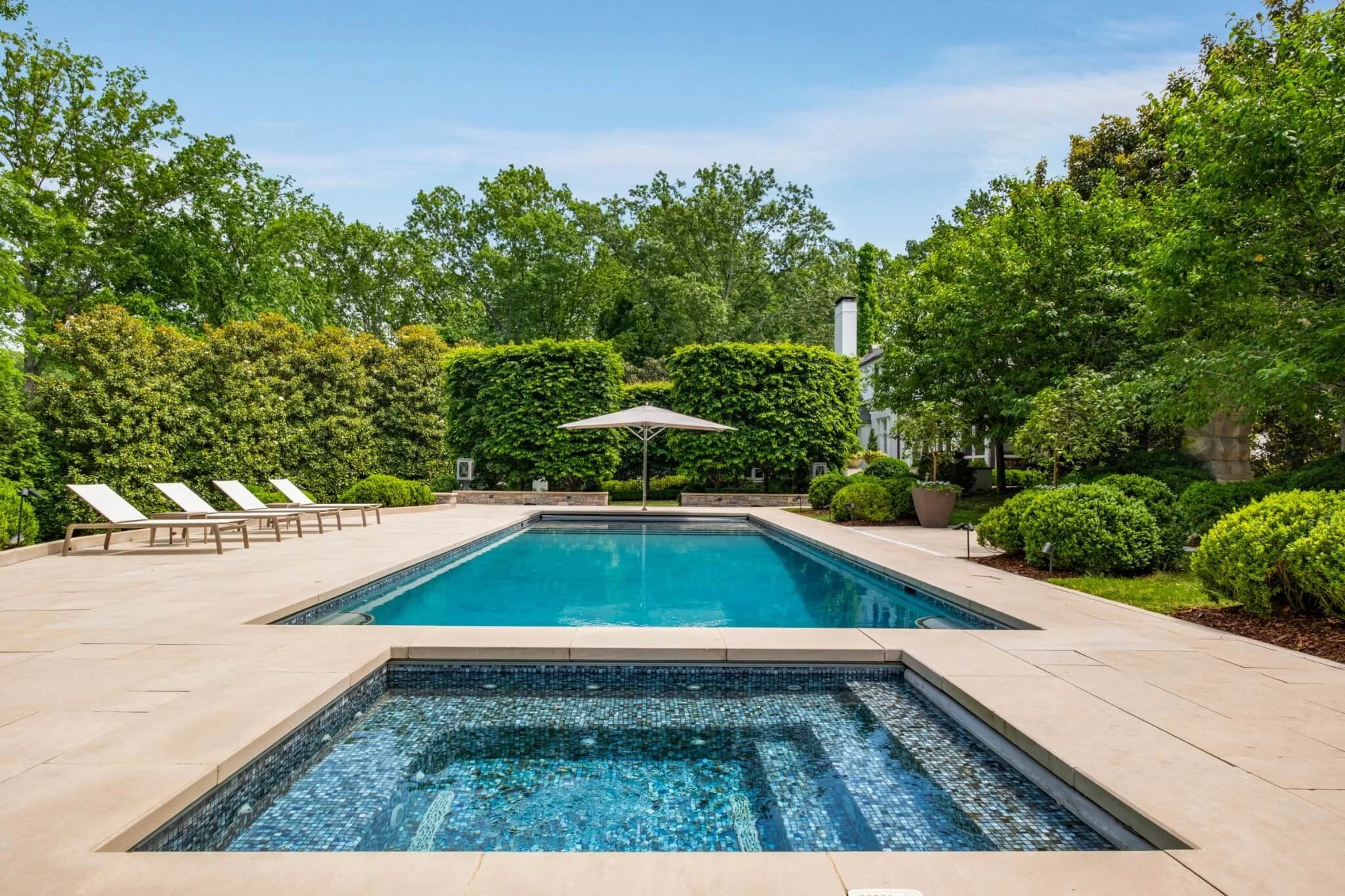 A backyard pool area with four lounge chairs and an umbrella, surrounded by lush green trees and shrubs, with a white house in the background under a partly cloudy sky.