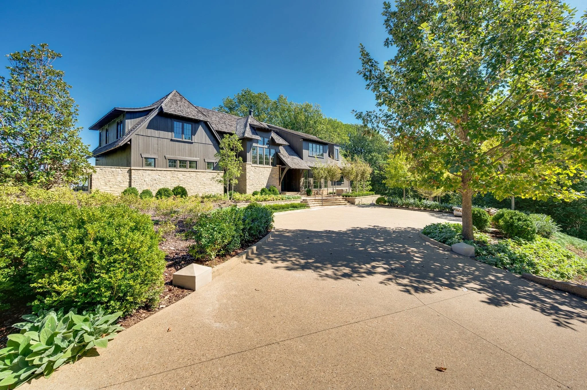 Large modern house with dark wood siding, surrounded by shrubs and trees, with a wide paved walkway leading up to the entrance under a blue sky.